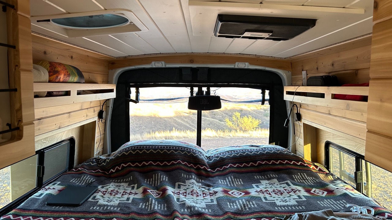 Inside a cozy rv rental: a comfy bed with patterned blanket, wood paneling, shelves, and a JBL speaker, overlooking scenic hills.. Ford Transit 2023