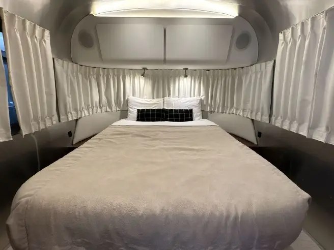 A serene RV rental bedroom features a beige bed, white pillows, and elegant pleated curtains lining the curved silver interior.. Airstream Globetrotter 2019