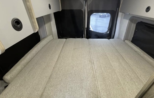 Relax in this RV rental's cozy camper van interior: a segmented bed, overhead storage, and a rear window view.. Winnebago Solis 2025