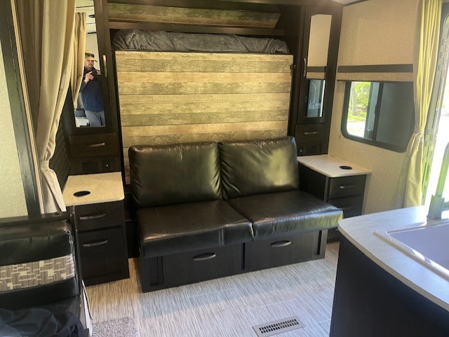 Cozy RV rental interior with black couch, pull-down bed, and side tables. A person is reflected in the mirror.. Forest River Surveyor Legend 2020