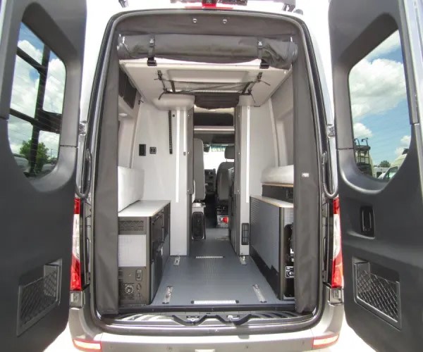 View from open rear doors into a grey camper van interior. Modular units, a central aisle, and overhead storage, ideal for an rv rental.. Winnebago Revel 2025