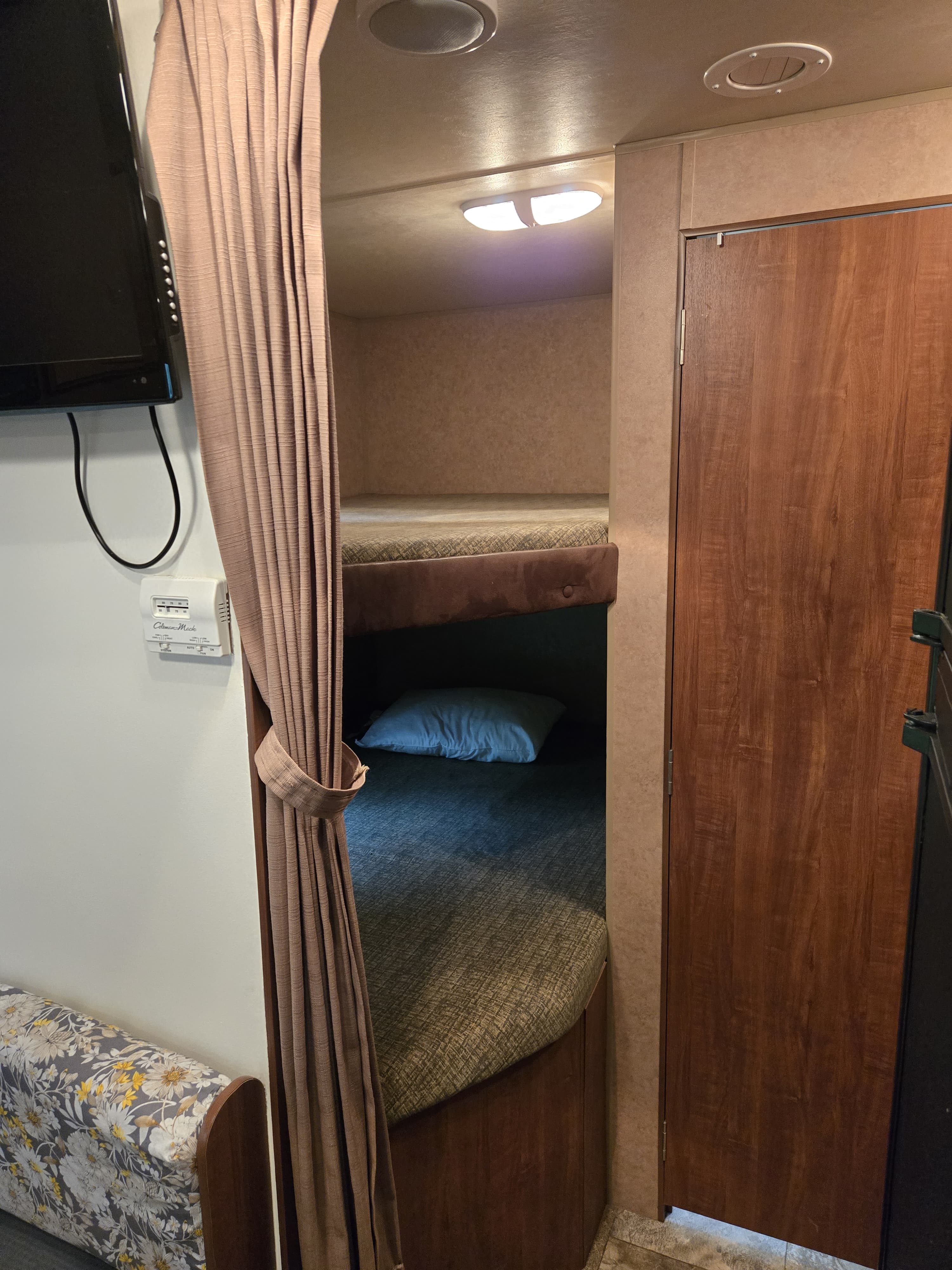 This rv rental features a bunk bed area with two beds, separated by a brown curtain. A TV and thermostat are on the left wall.. Jayco Jay Flight 2013