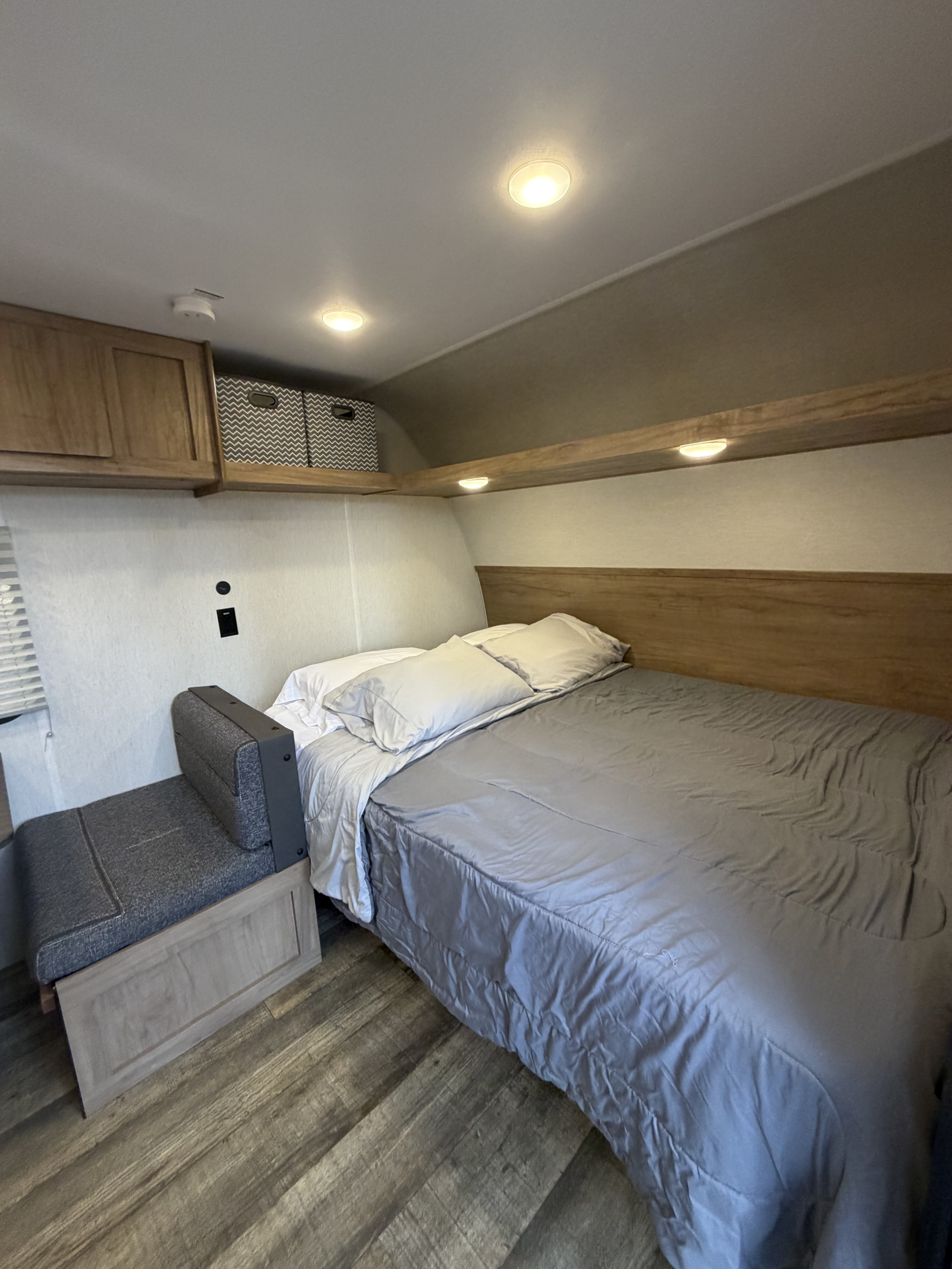 Cozy RV rental bedroom with a bed, gray bedding, small bench seat, overhead storage, and warm lighting.. Forest River Cherokee Wolf Den 2024
