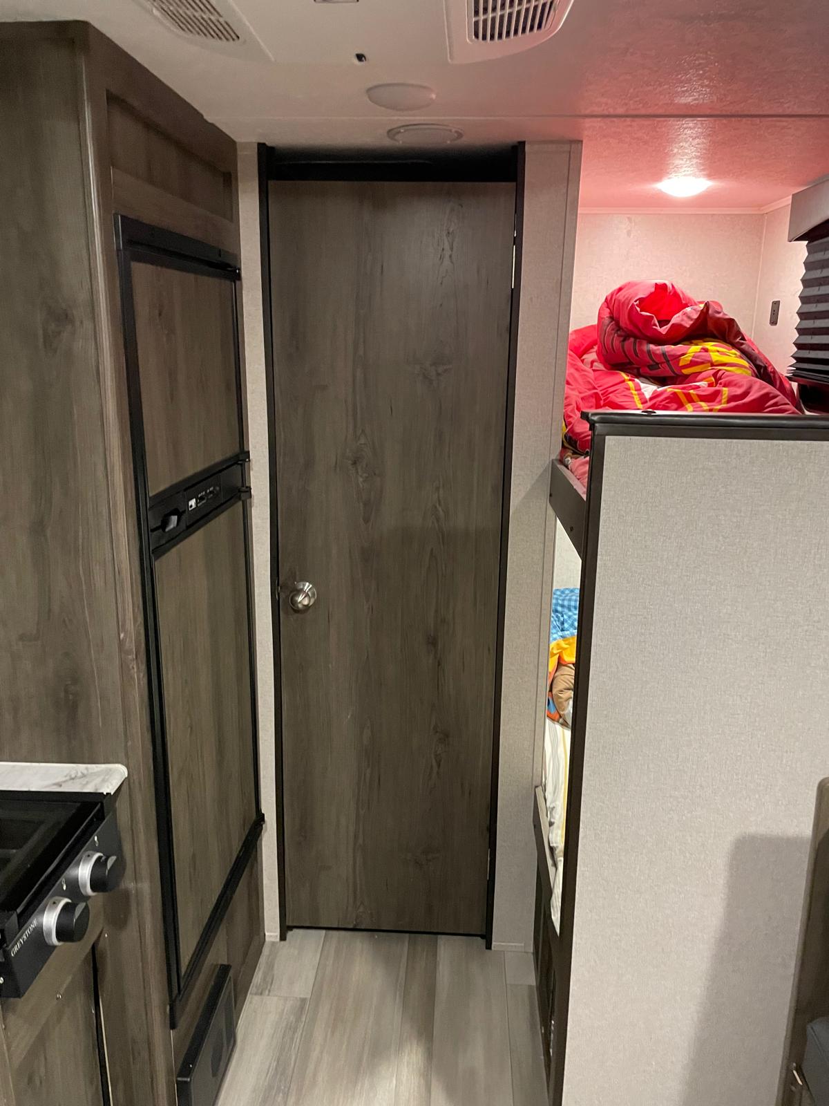 Inside this RV rental, featuring a compact kitchenette, an interior door, and cozy bunk beds with a bright red comforter.. Coachmen Viking 17BHS 2021