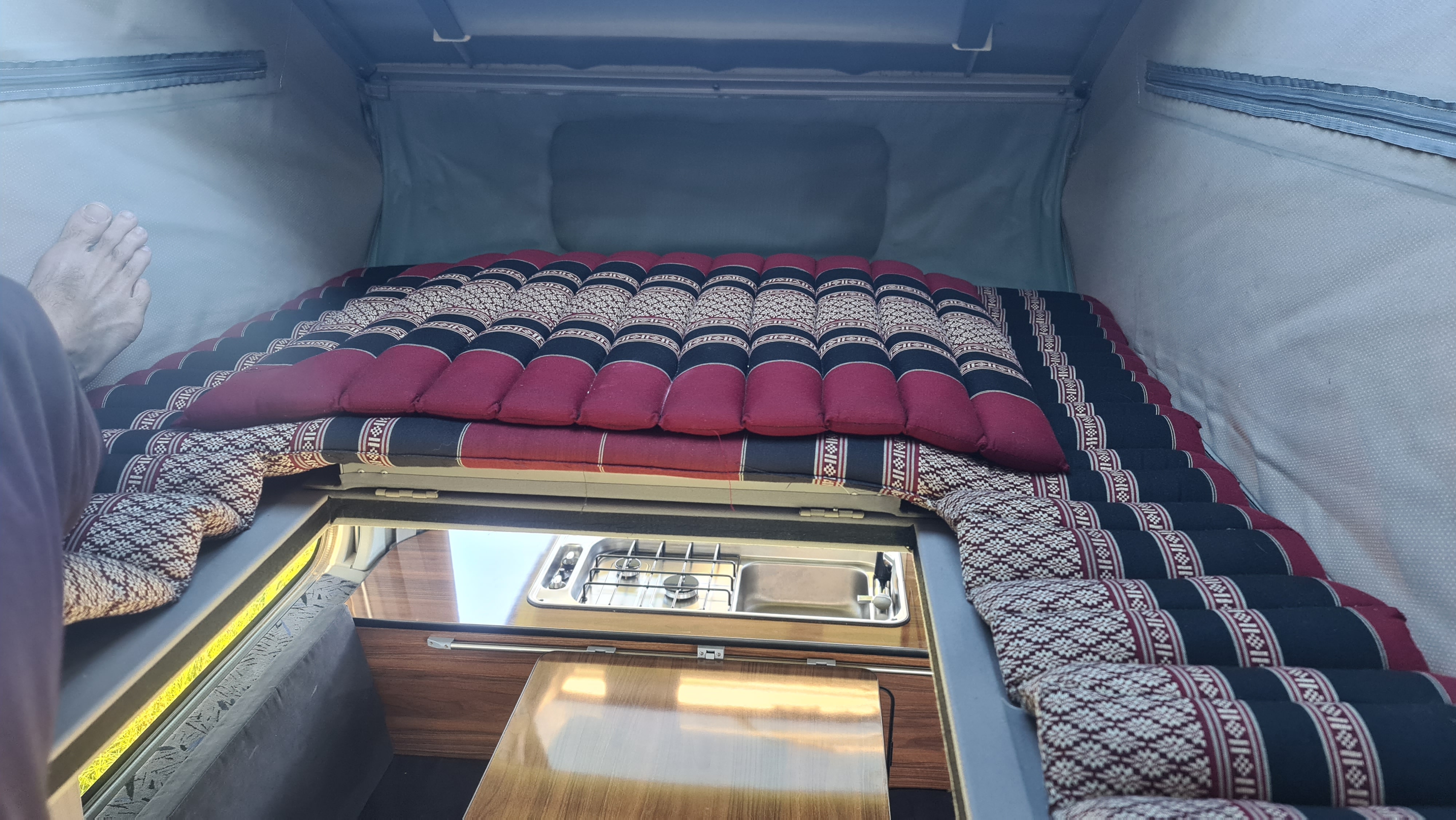 Cozy RV rental interior: patterned sleeping area above a compact kitchen with stove/sink. Pop-top camper for adventurers.. Mazda Bongo 2002