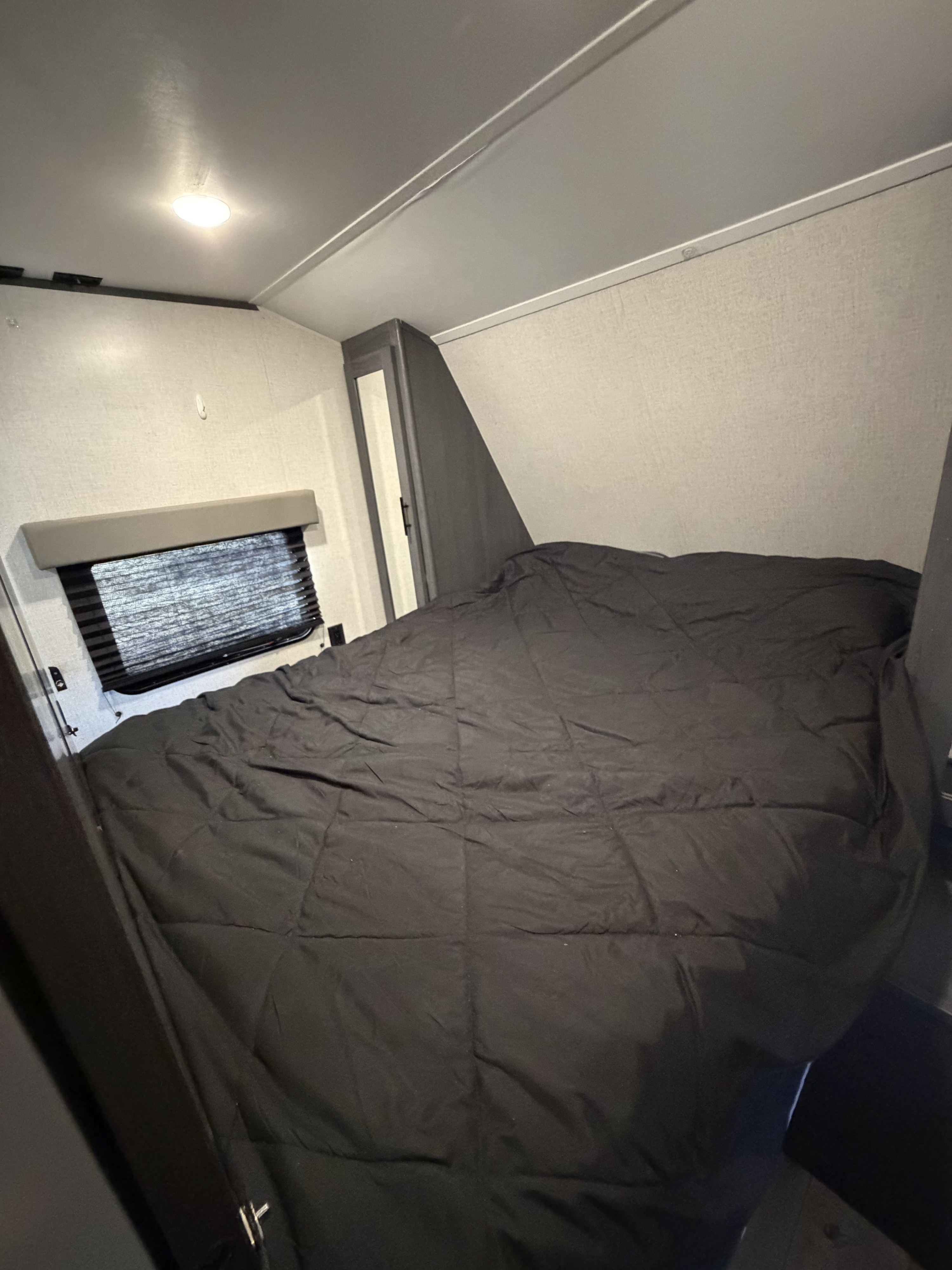 A bedroom in an RV rental, showing a bed with a black comforter, a window with blinds, and a built-in cabinet.. Dutchmen Aspen Trail LE 26BH 2022