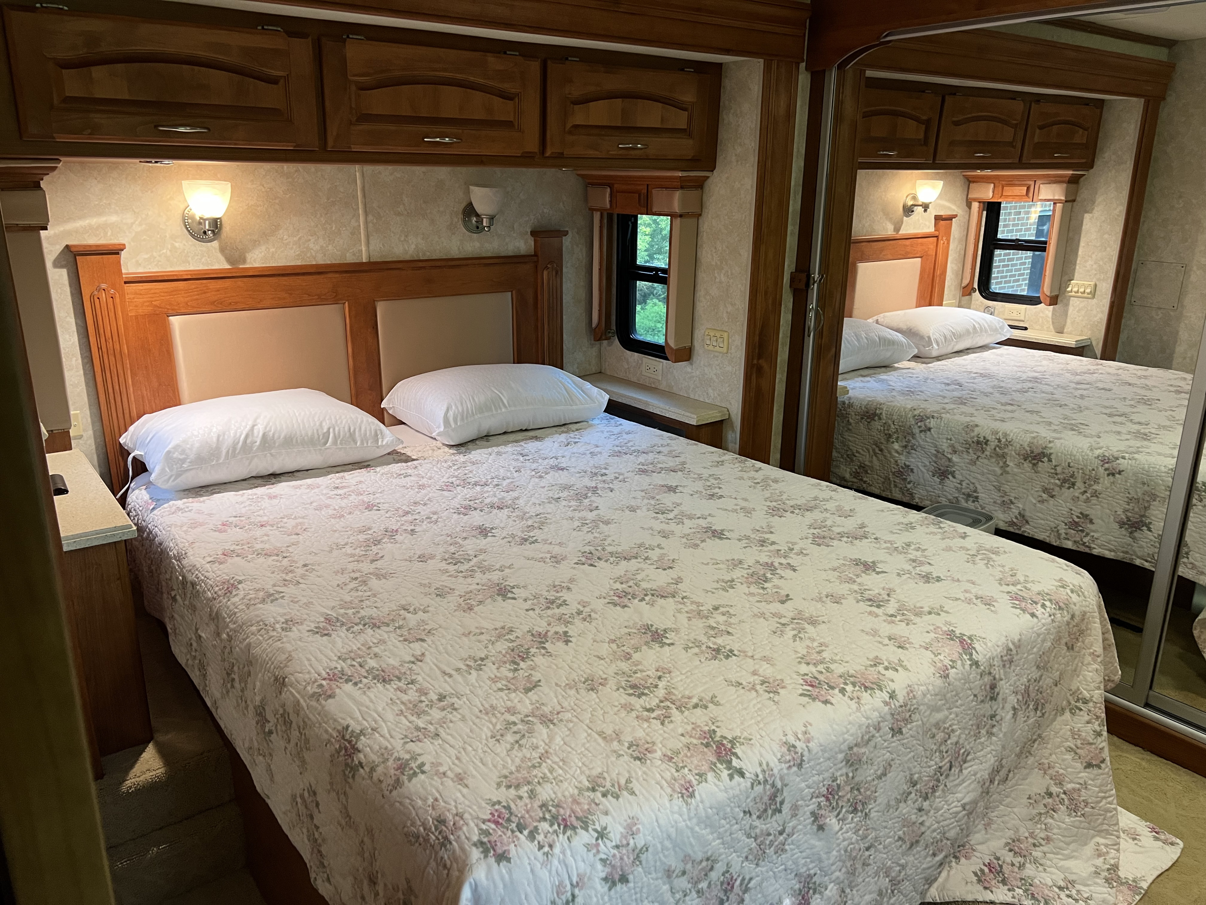 Cozy RV rental bedroom: bed with floral comforter & white pillows, wooden cabinets, wall lights, and mirror.. Holiday Rambler Endeavor 2007