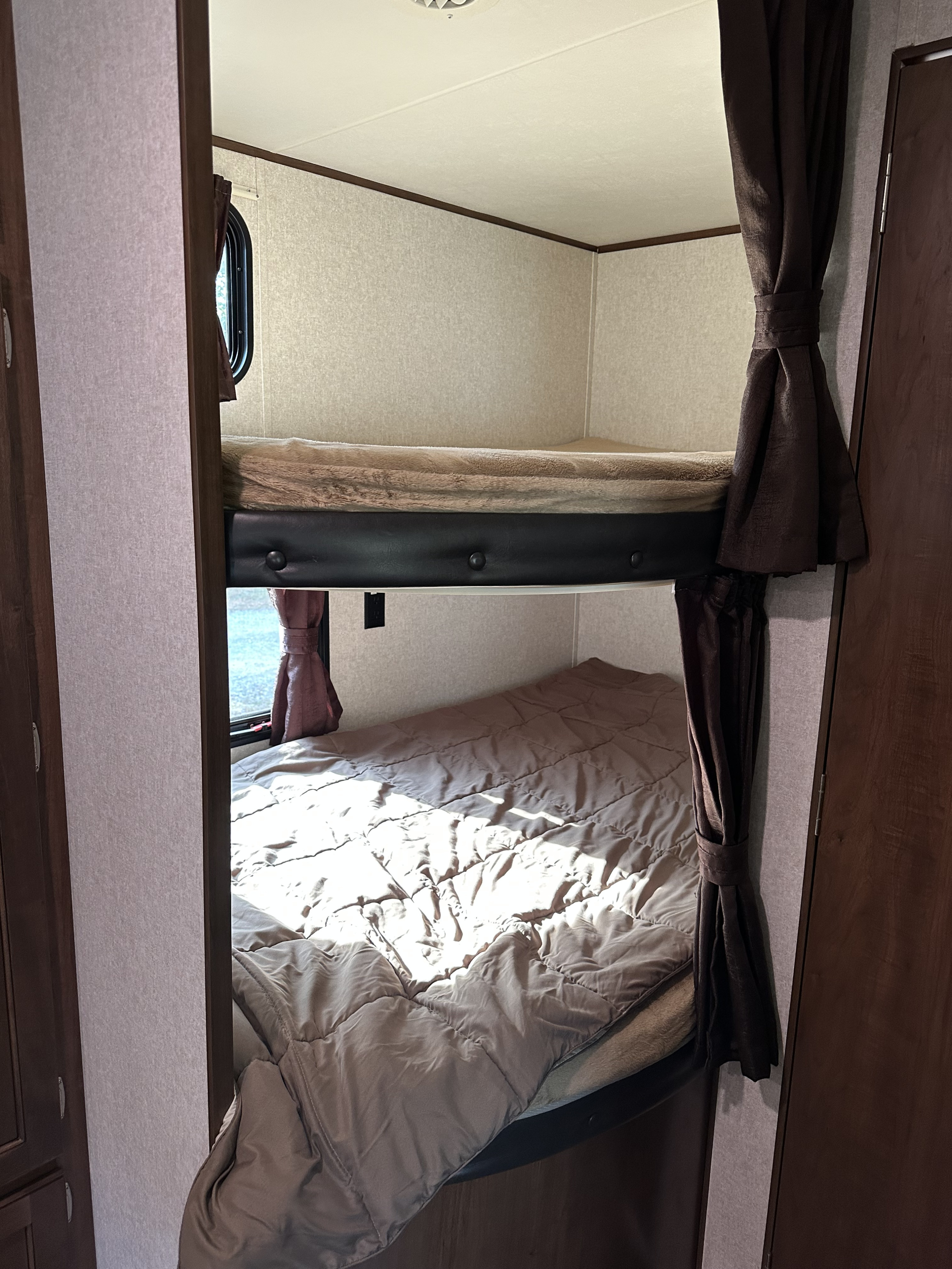 Cozy RV rental bunk beds with warm bedding. Sunlight illuminates the bottom bunk, framed by dark brown tied-back curtains.. Jayco Jay Flight SLX 2017