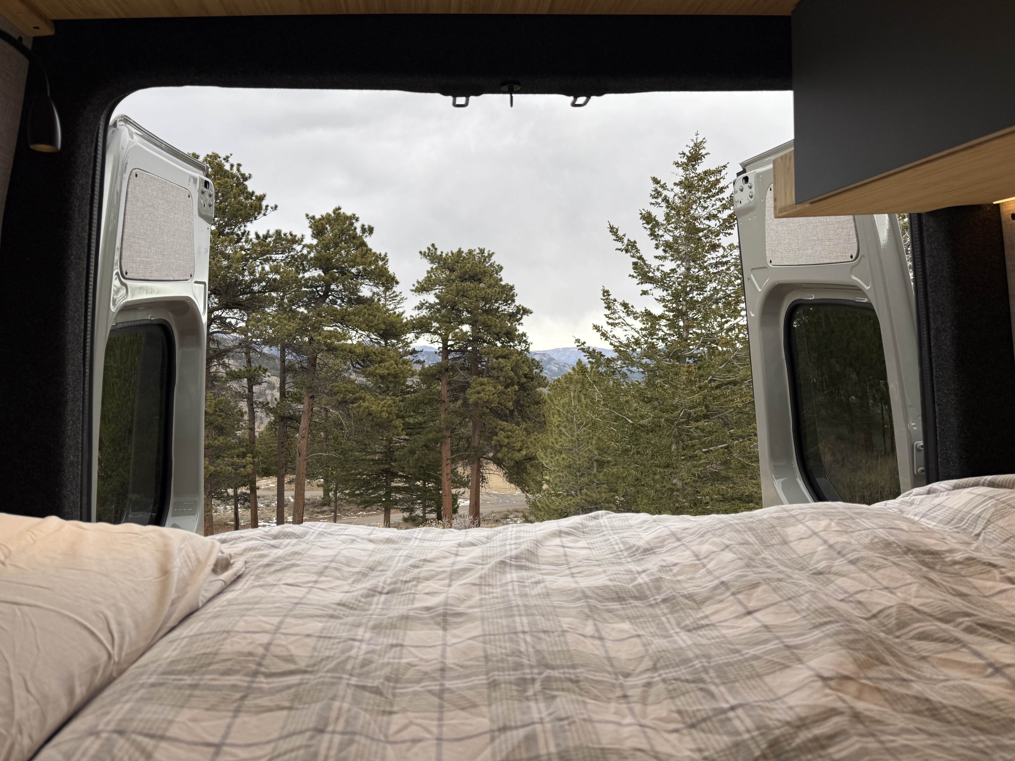 Wake up to nature! This RV rental bed with plaid bedding offers a serene view of pine trees, mountains, and a cloudy sky from open van doors.. Mercedes-Benz Sprinter 2026