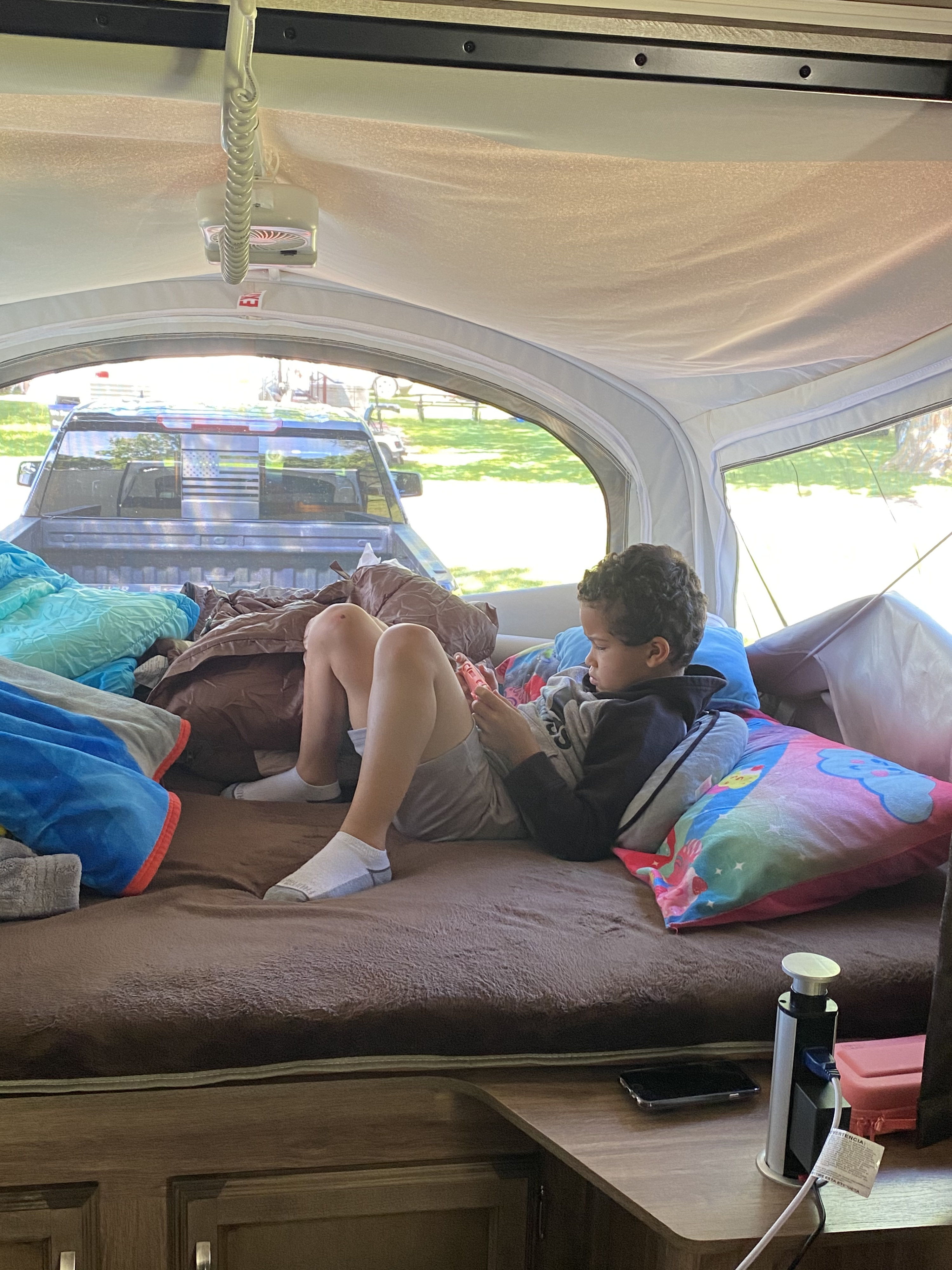Child relaxing in an RV rental pop-up camper, playing a handheld game. A truck and outdoor scenery are visible outside.. Jayco Jay Feather 2020