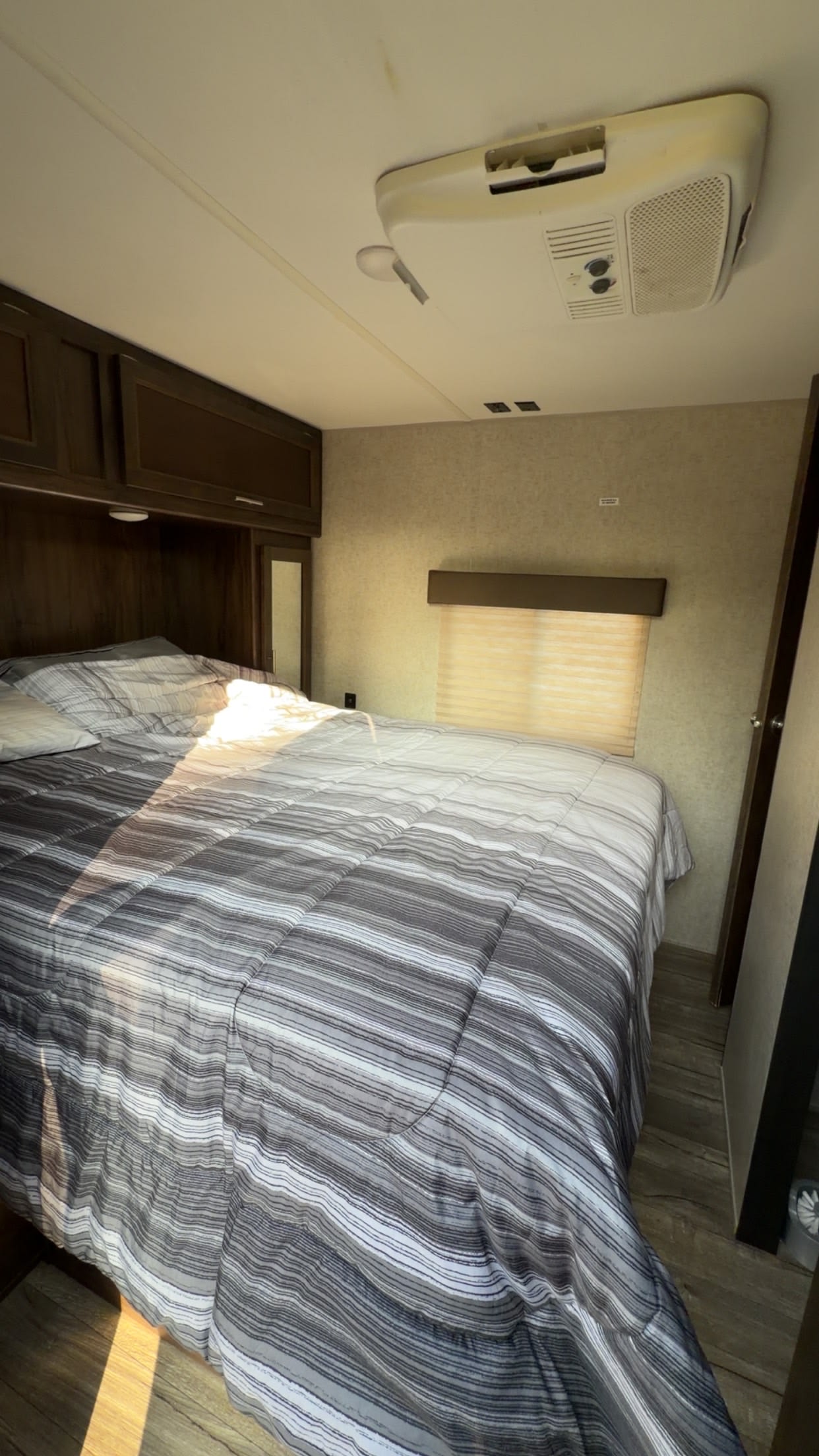 This RV rental bedroom features a comfy bed with striped bedding, overhead cabinets, and ceiling AC. Sunlight streams in.. Forest River Cherokee 2018