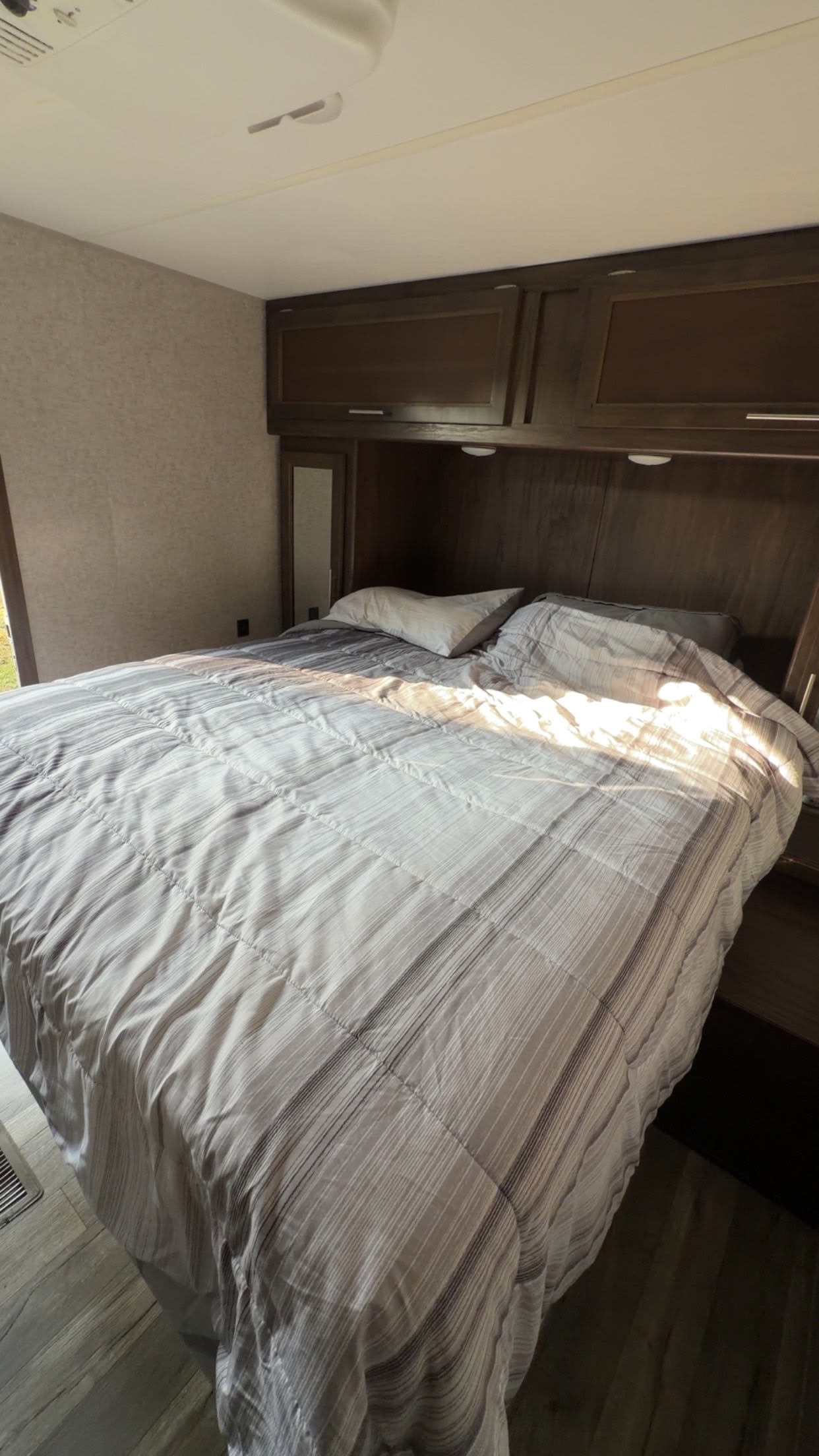 This RV rental bedroom offers a comfortable bed with a striped comforter, overhead wood cabinetry, and a mirror, illuminated by sunlight.. Forest River Cherokee 2018