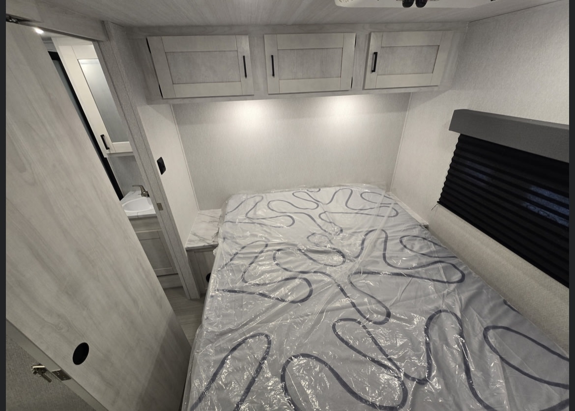Bright RV rental bedroom featuring a bed, overhead cabinets, and a visible bathroom sink. Ready for your travels!. East to West Entrada 2025