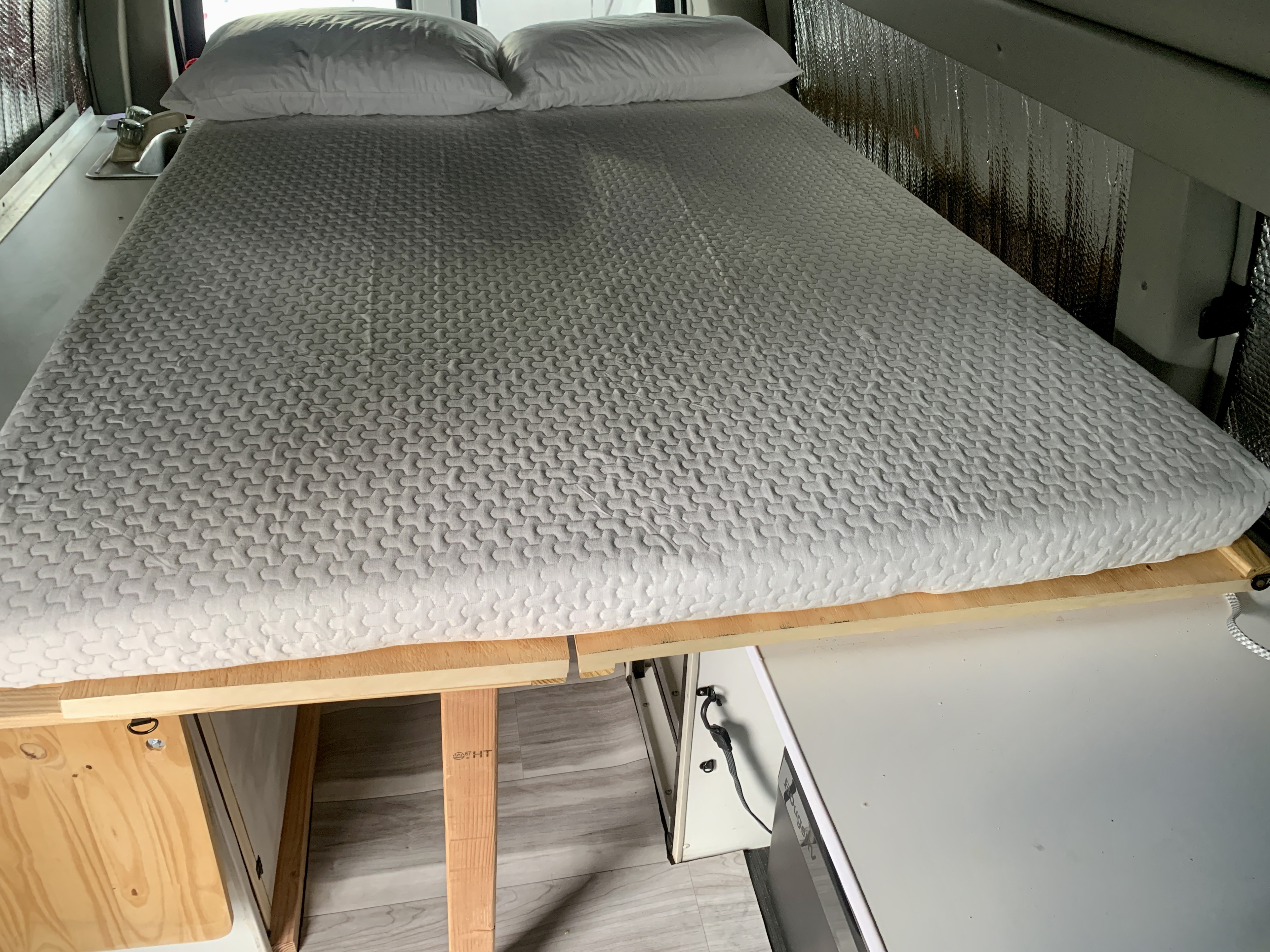 Cozy bed and sink area inside an RV rental. Features a white quilted mattress, pillows, a wooden frame, and insulated walls.. Chevrolet Express 2011