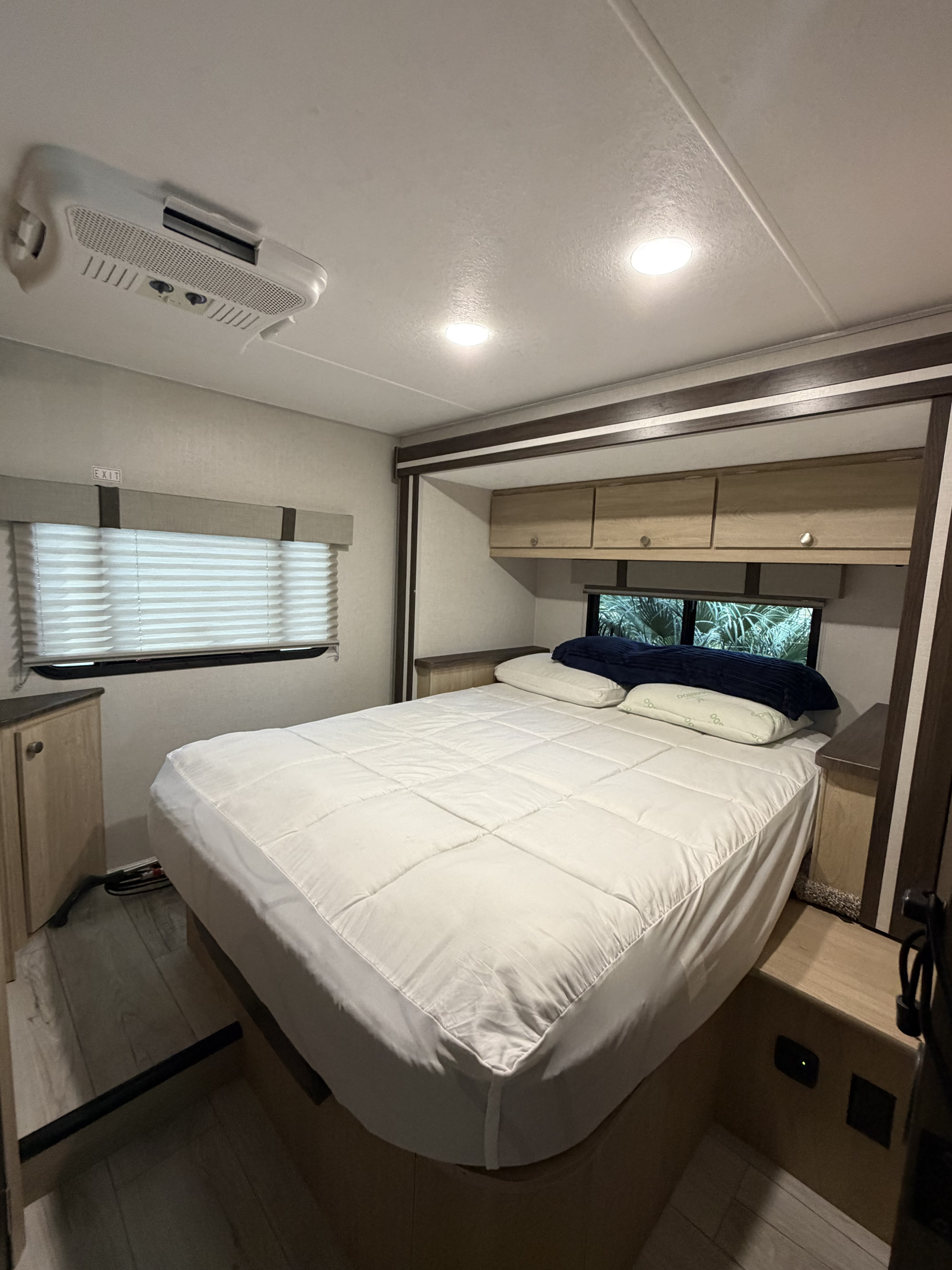 This RV rental bedroom features a cozy bed with white bedding, overhead storage, AC unit, and a window with blinds.. Coachmen Leprechaun 2025