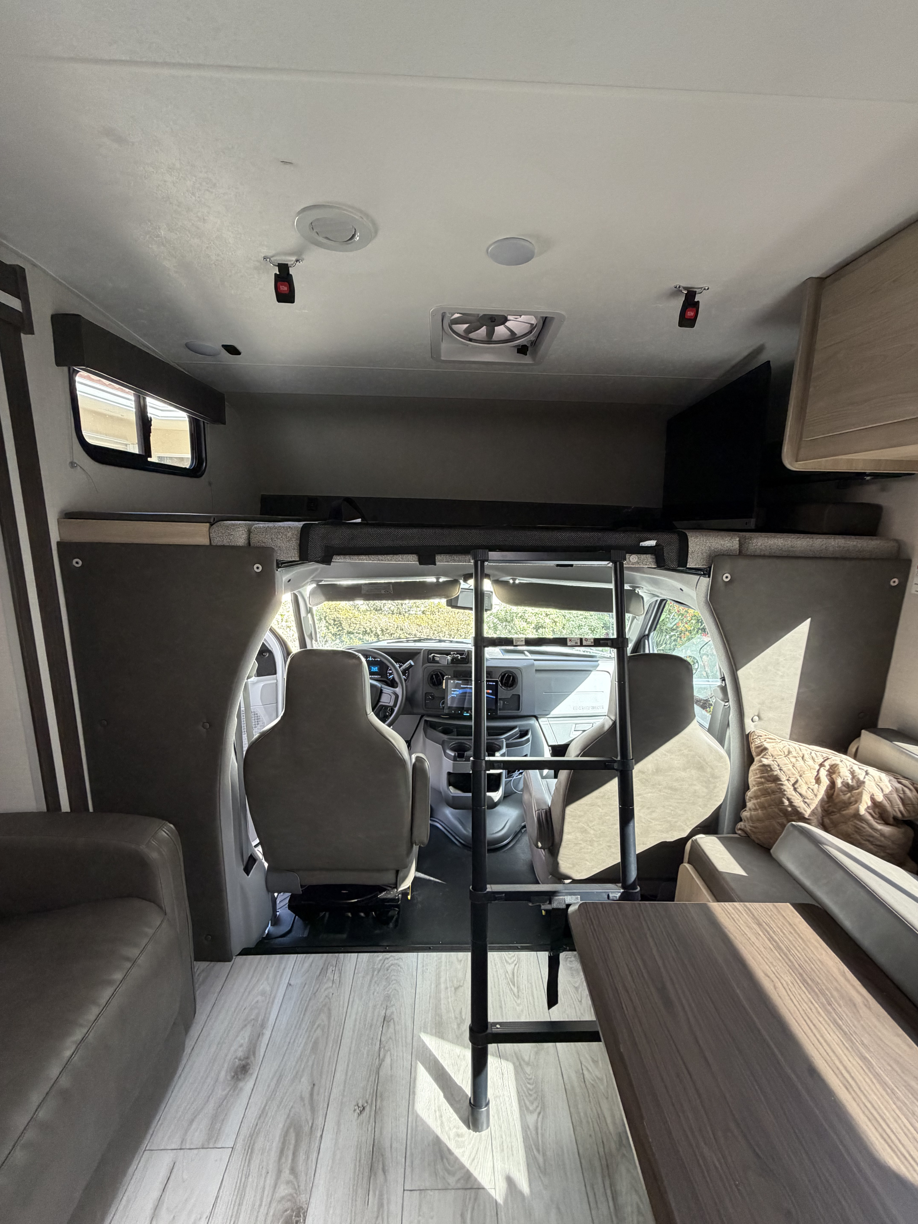 Interior of an rv rental looking forward: light wood floor, gray seating, ladder to over-cab bunk, cockpit, and table.. Coachmen Leprechaun 2025