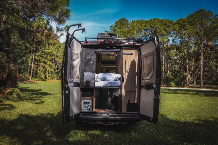 Open RV rental reveals a bed, storage, & controls inside, parked on green grass surrounded by tall trees.. Winnebago Travato 2022