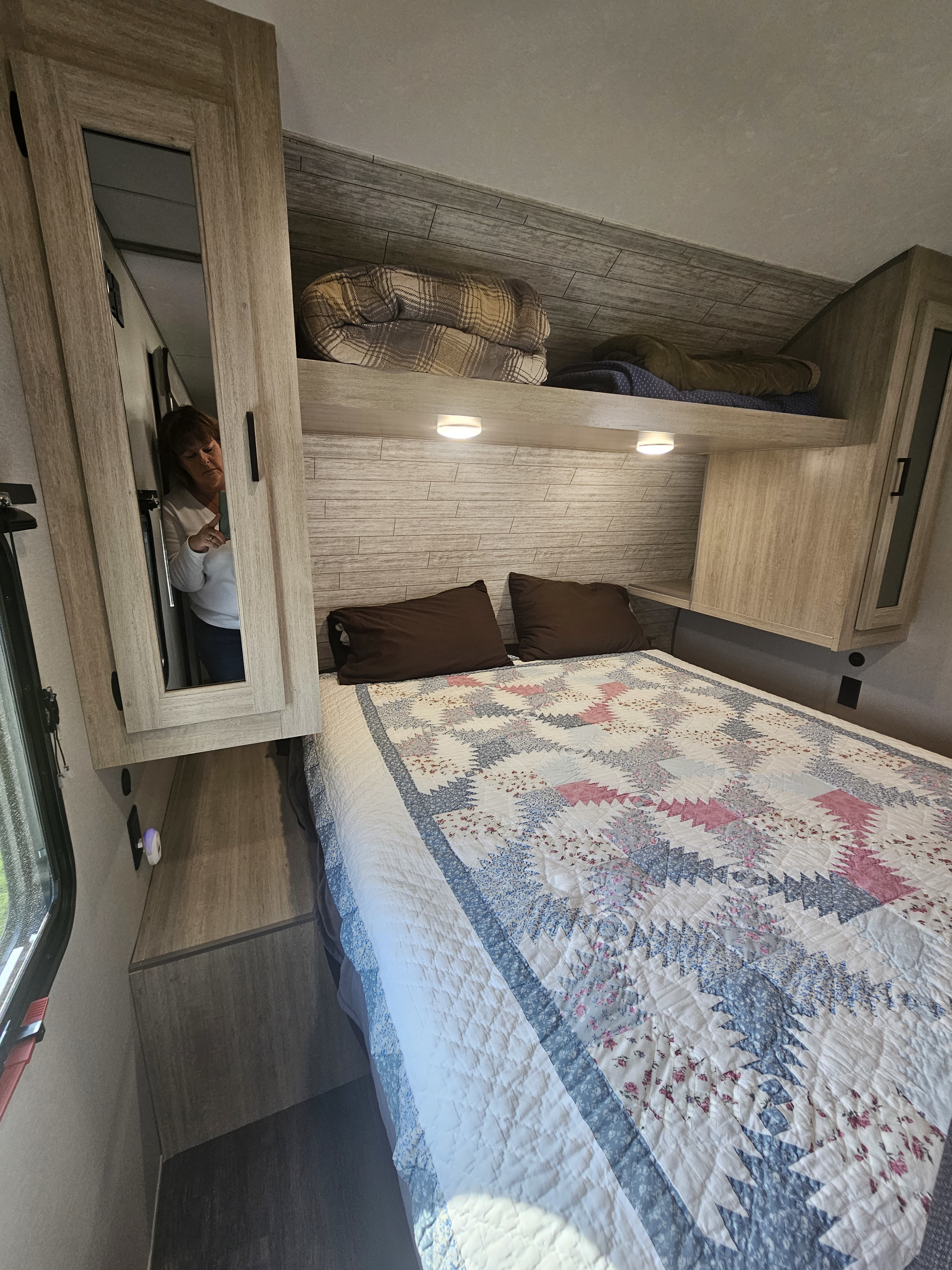 Cozy RV rental bedroom with a patterned quilt, dark pillows, ample overhead storage, and a person's reflection.. Forest River Aurora Aurora 2026
