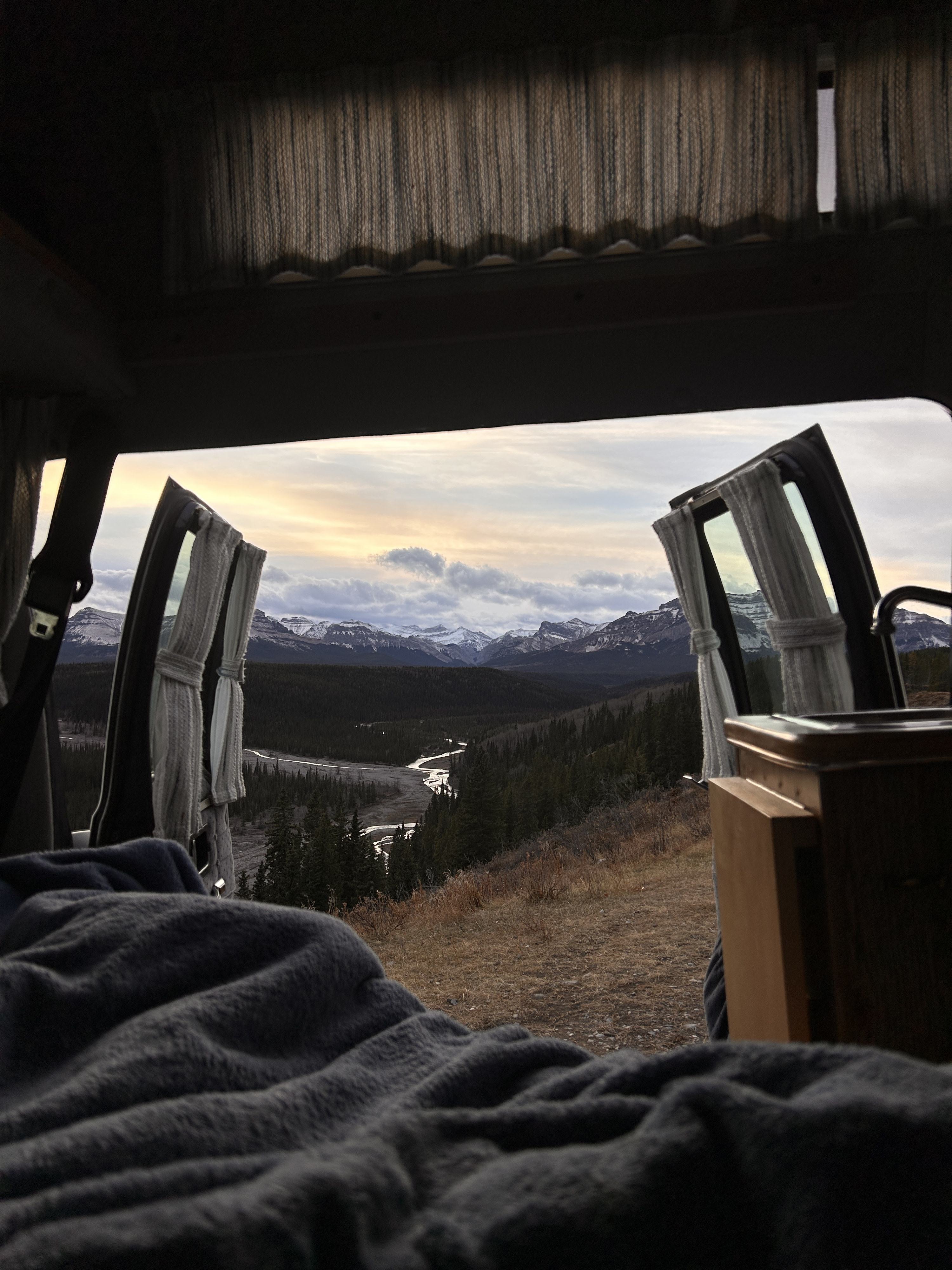 Waking up in an RV rental to a breathtaking mountain view, winding river, and forest. Cozy blanket in foreground.. Dodge B Van 1987