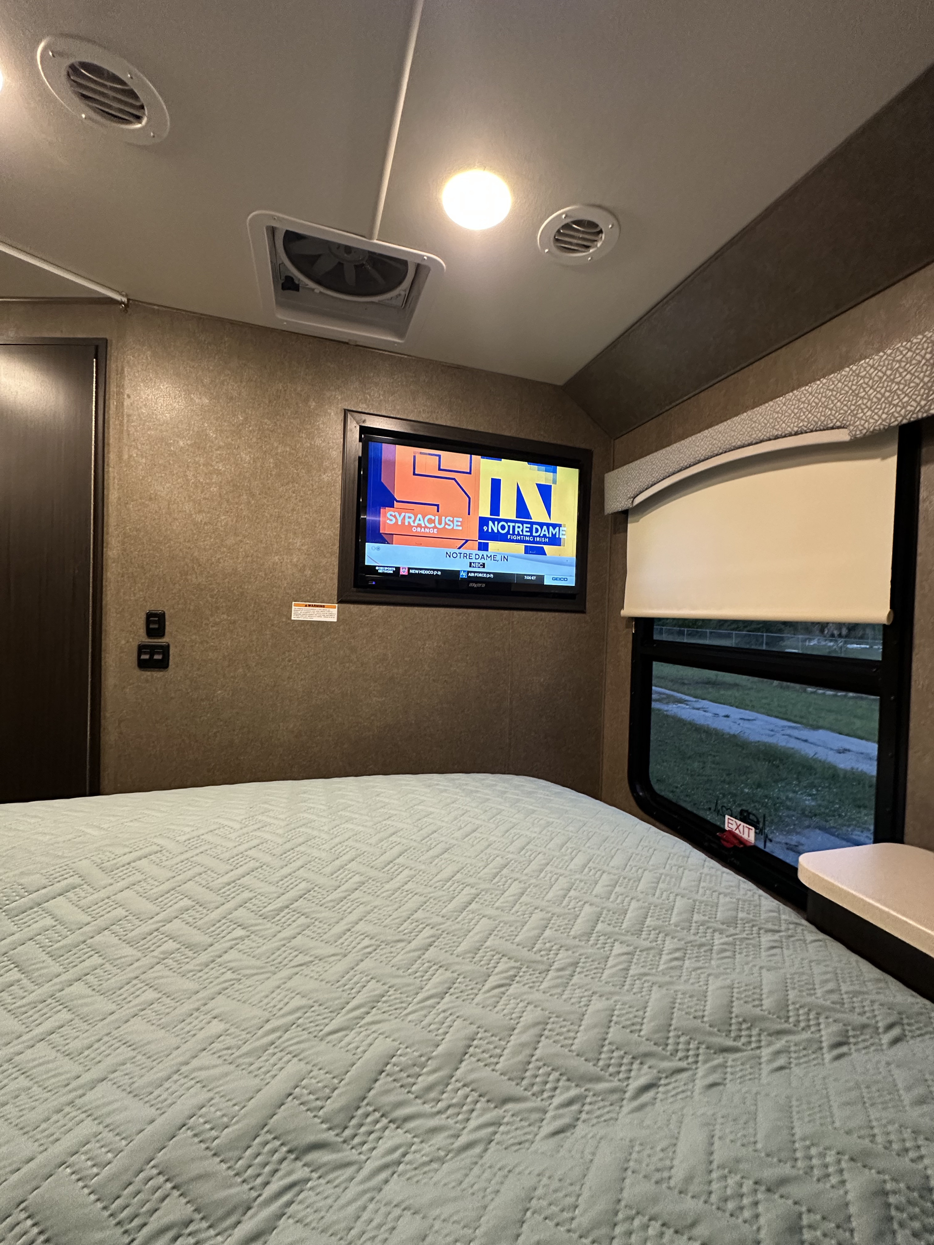 RV rental bedroom interior: A TV displays a Syracuse vs. Notre Dame game above a light blue quilted bed. Window has an EXIT sign.. Thor Vegas 2018