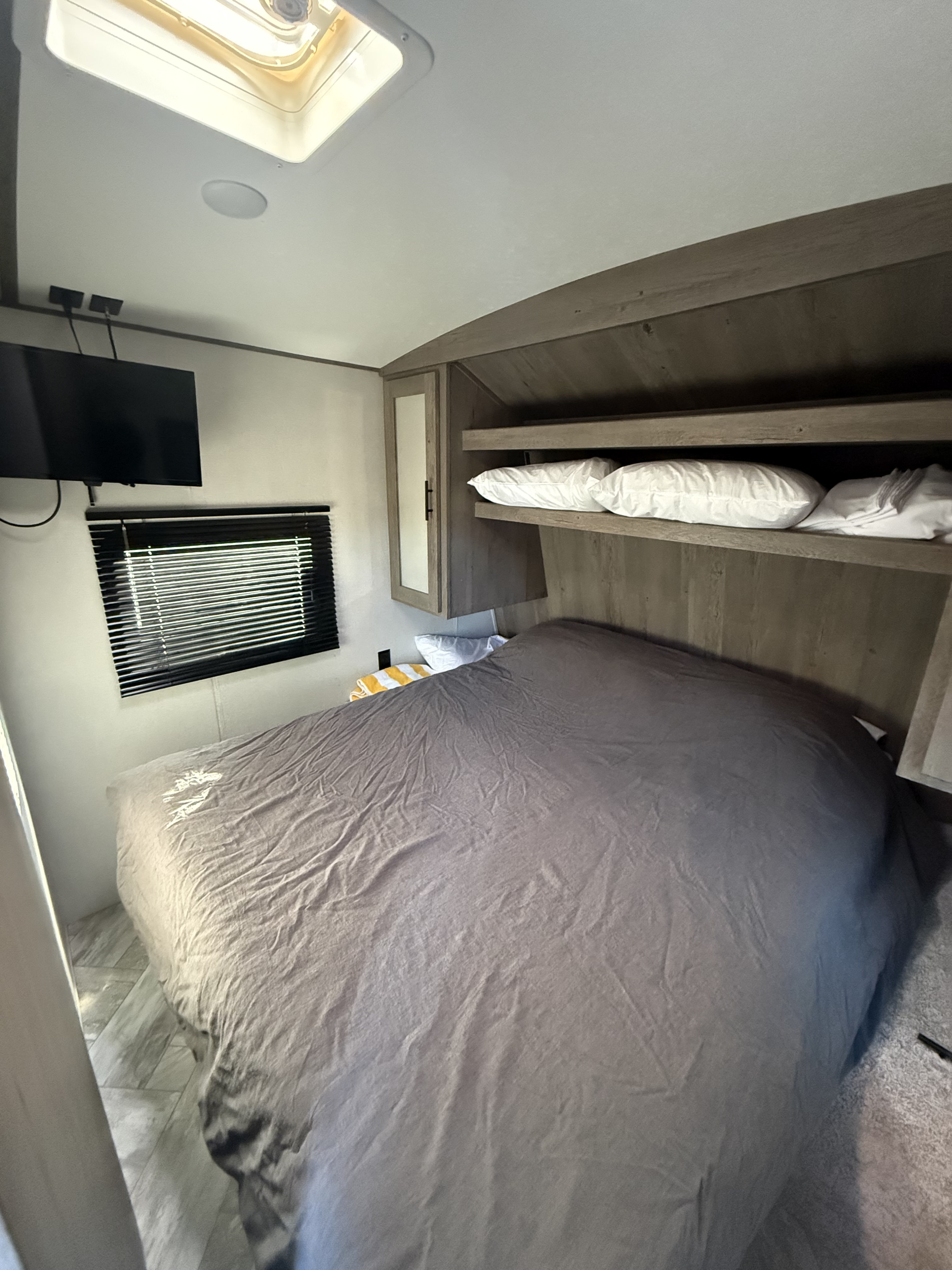 Cozy RV rental bedroom featuring a bed with gray linens, wall-mounted TV, window, skylight, and wooden storage.. Keystone RV Springdale 2021