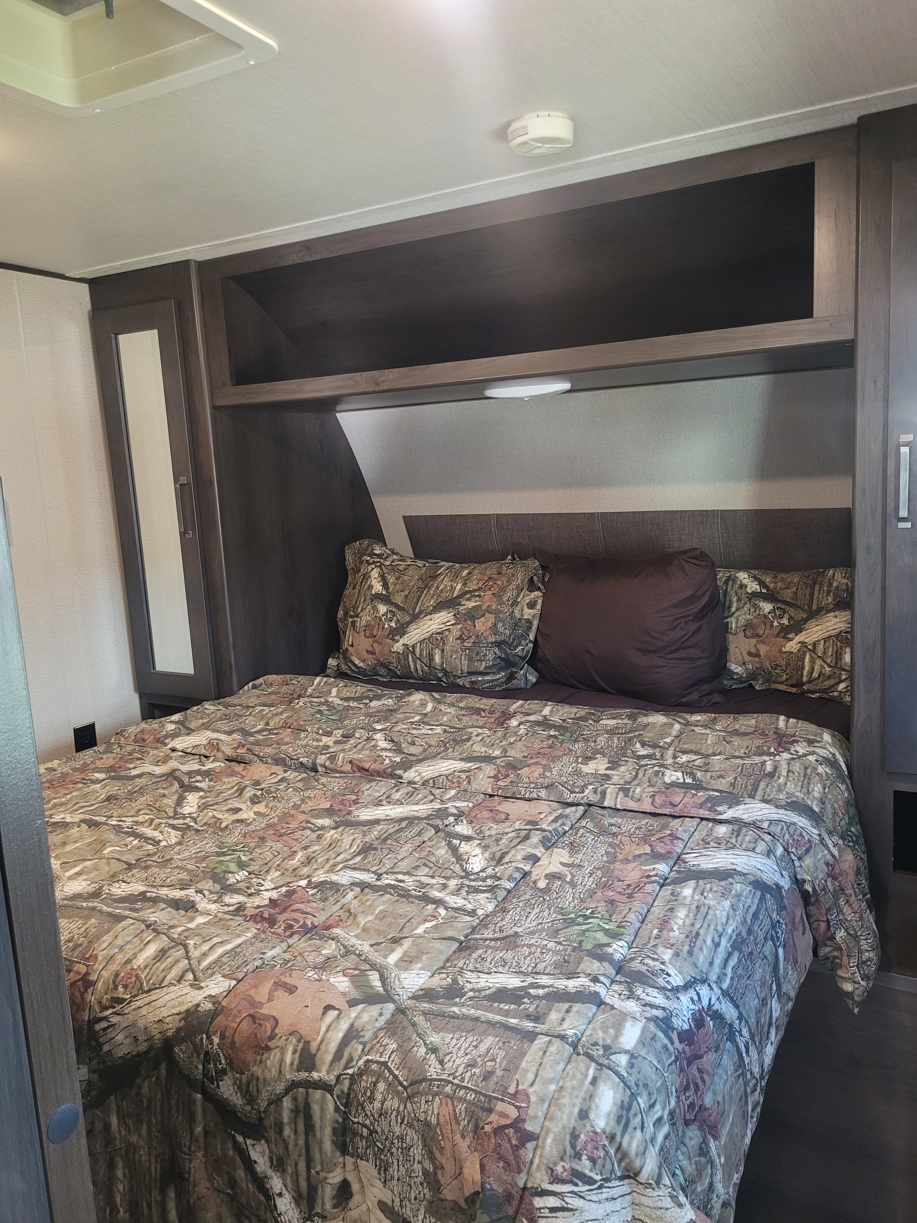 This RV rental bedroom features a bed with camouflage bedding, dark wood cabinets, a headboard, ceiling vent & smoke detector.. Grand Design Transcend Xplor 2021