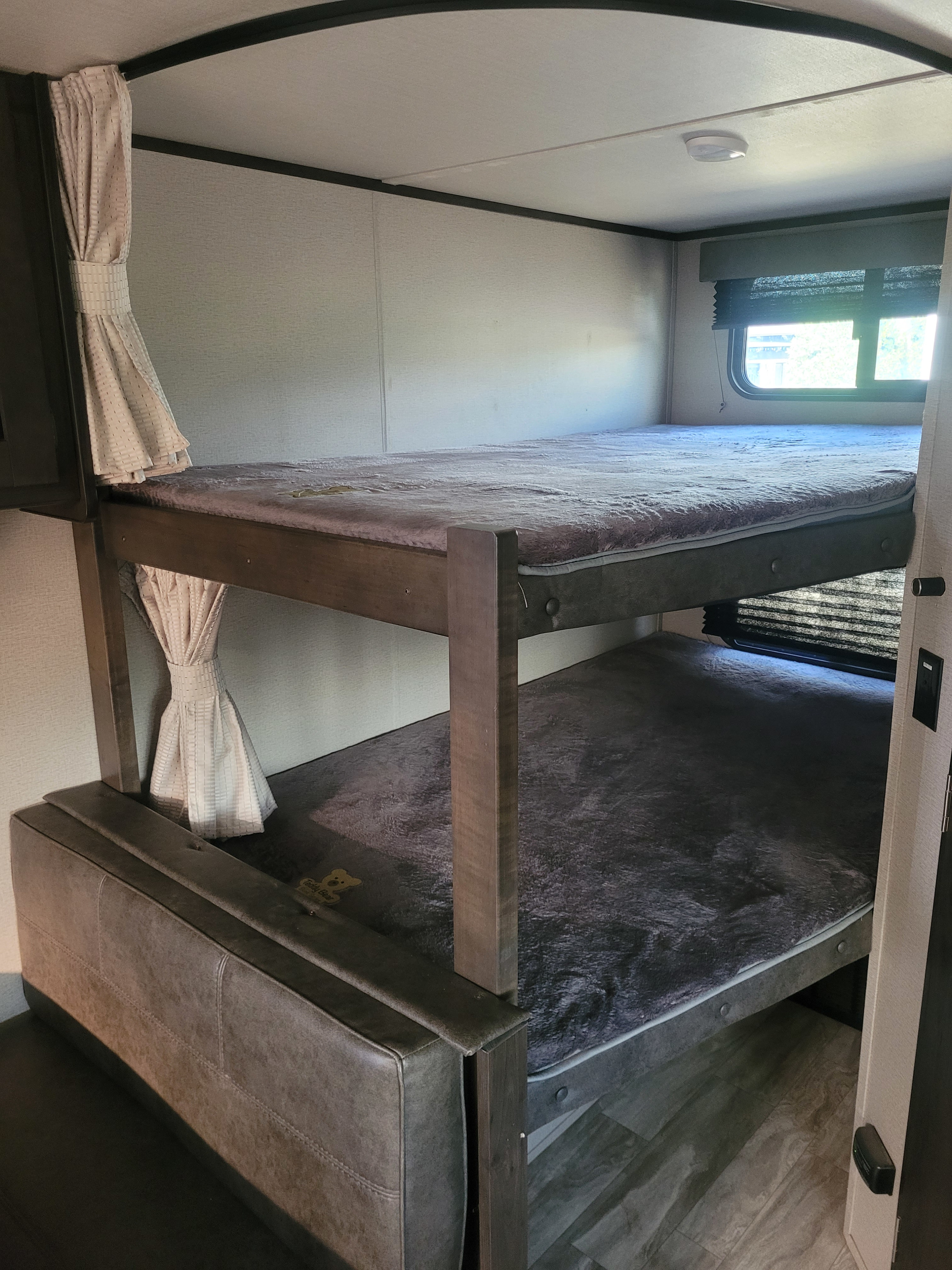 Inside an RV rental, this compact sleeping area features wooden bunk beds with soft grey bedding, tied-back curtains, and a window.. Grand Design Transcend Xplor 2021