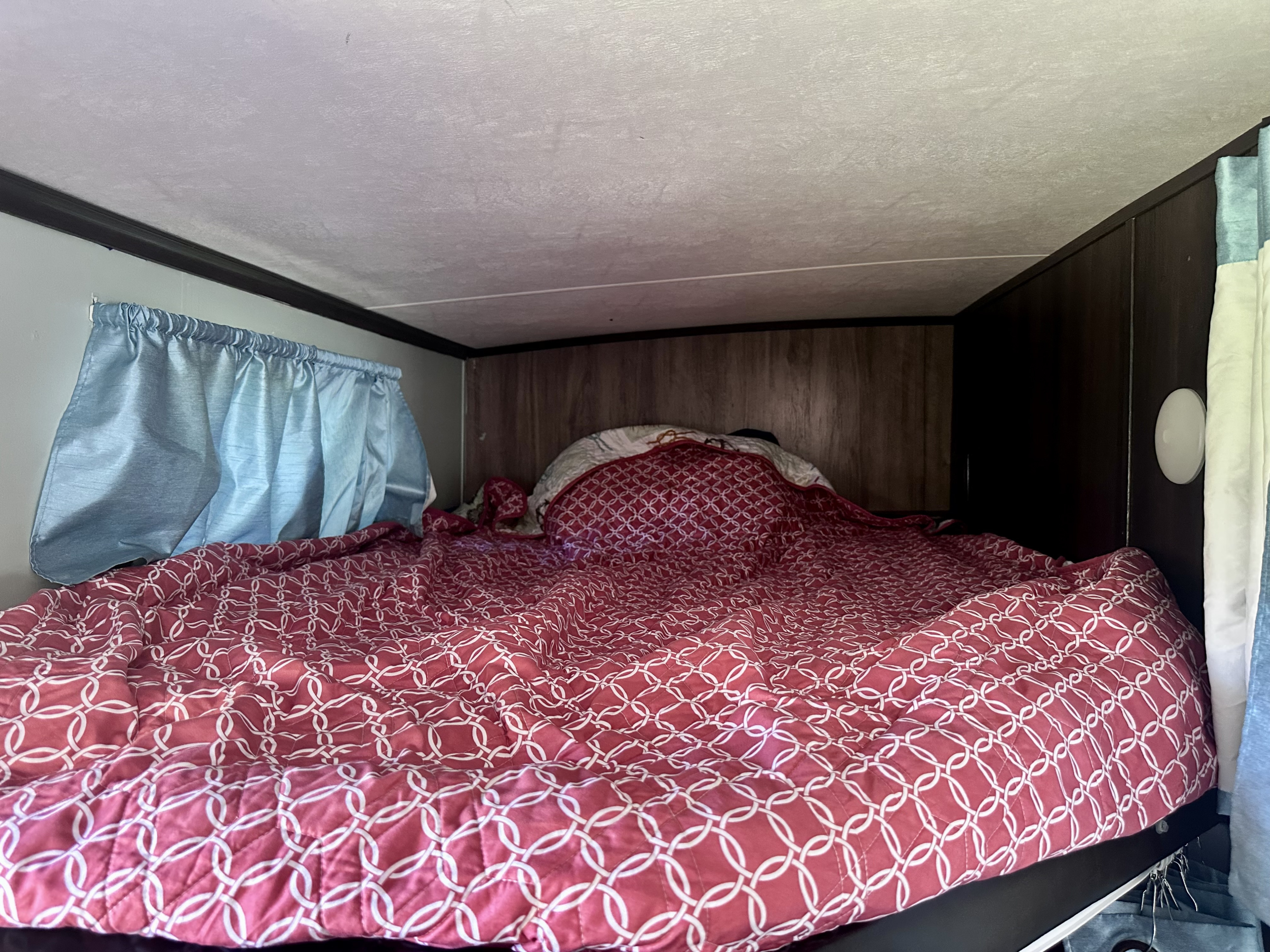 Cozy RV rental bunk bed with a red patterned comforter and blue curtains, ideal for a compact sleeping area.. Jayco White Hawk 2019