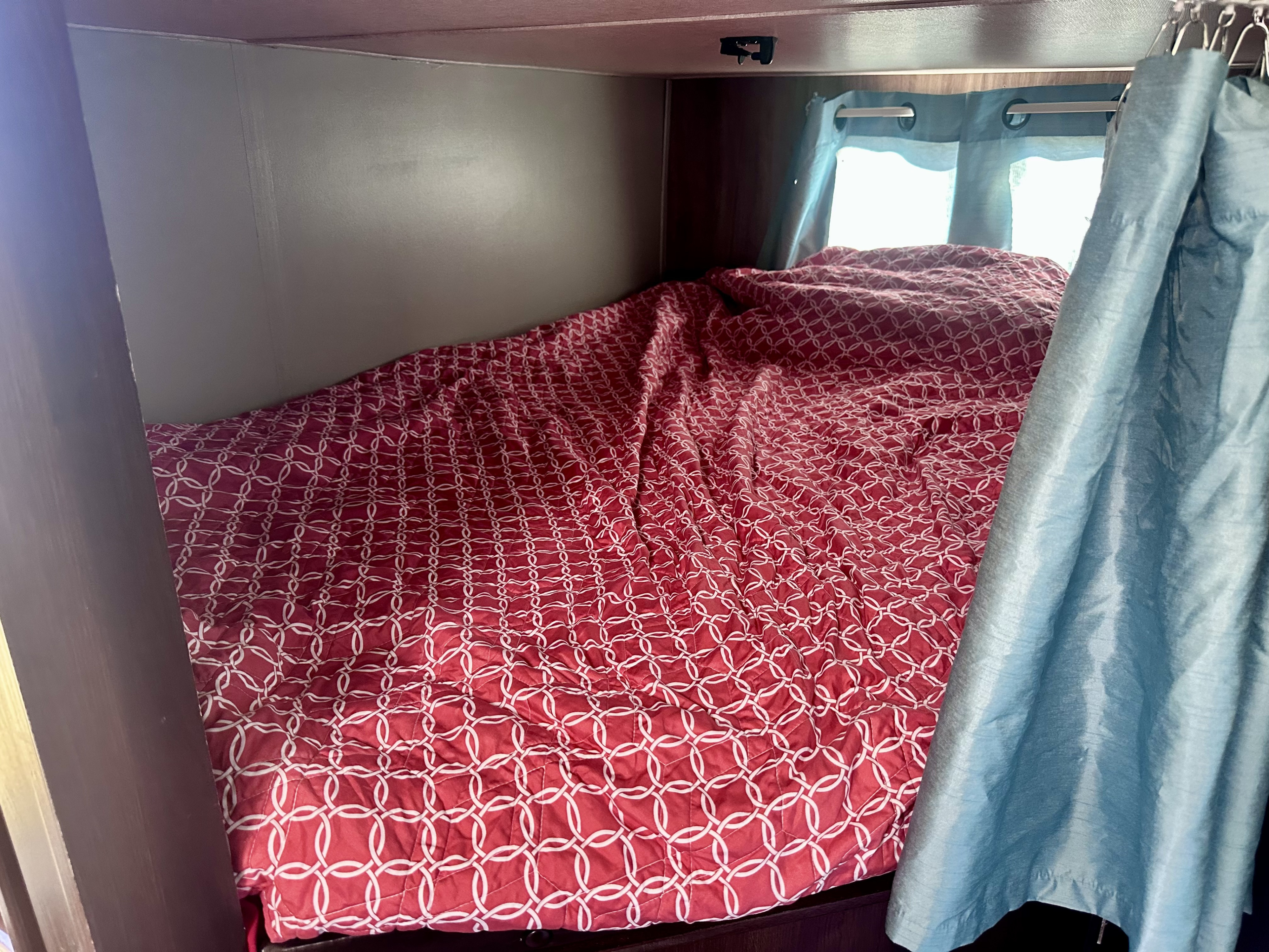 Cozy RV rental bunk bed with a red patterned comforter and light blue privacy curtain, ideal for compact travel.. Jayco White Hawk 2019