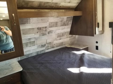 Inside an RV rental, a bedroom with a dark bed, rustic wood accent wall, and overhead storage.. Dutchmen Coleman Lantern 2021