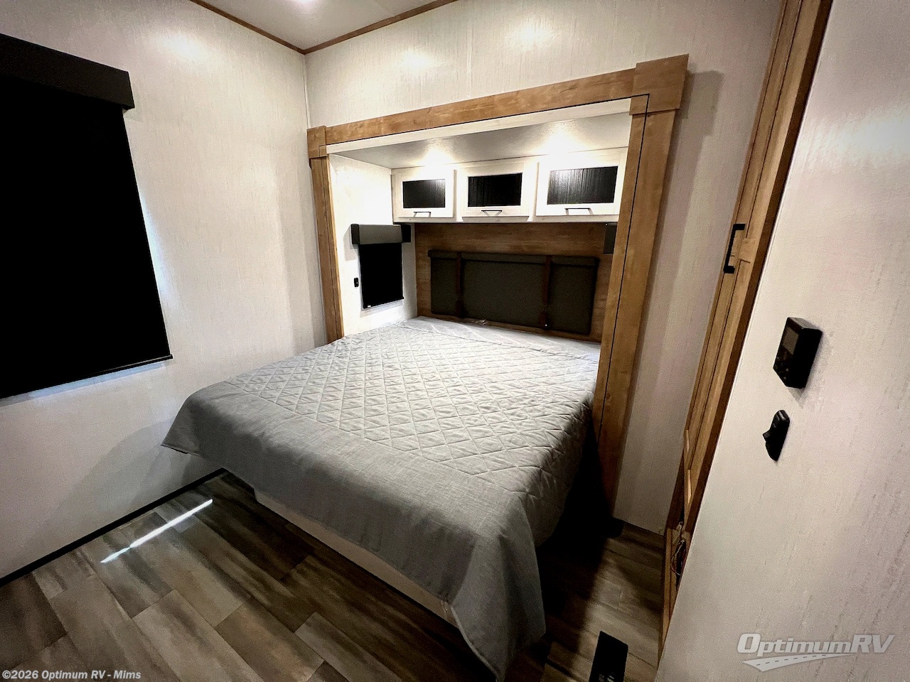 Comfortable RV rental bedroom with a quilted bed, wood-framed headboard, overhead cabinets, and a dark window shade.. Heartland RVs 321FL 2023