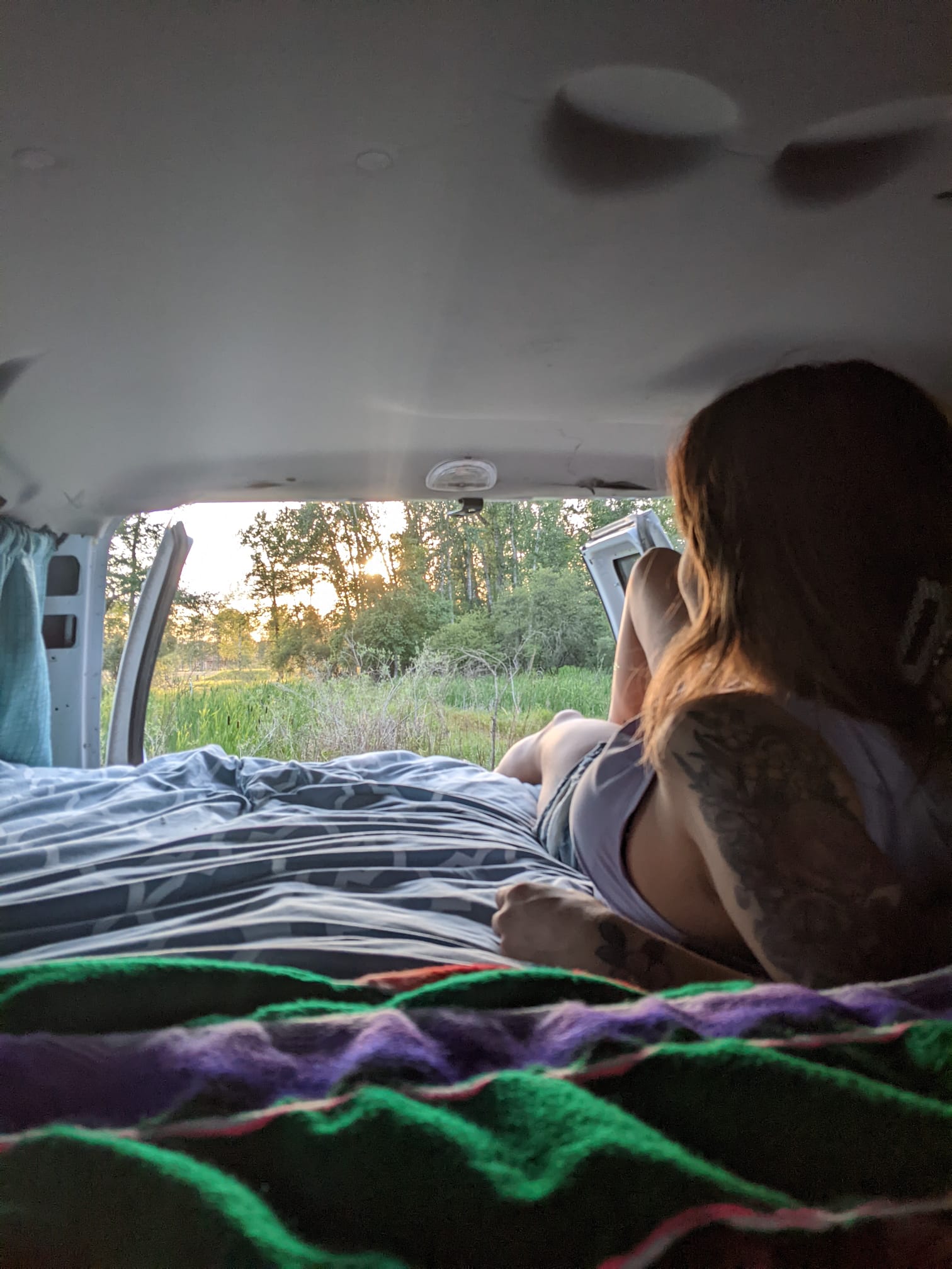 Relaxing in an RV rental, a woman enjoys the peaceful nature view from her cozy bed at sunset. #vanlife #travel. Glacier Campers Glamper Van 2014