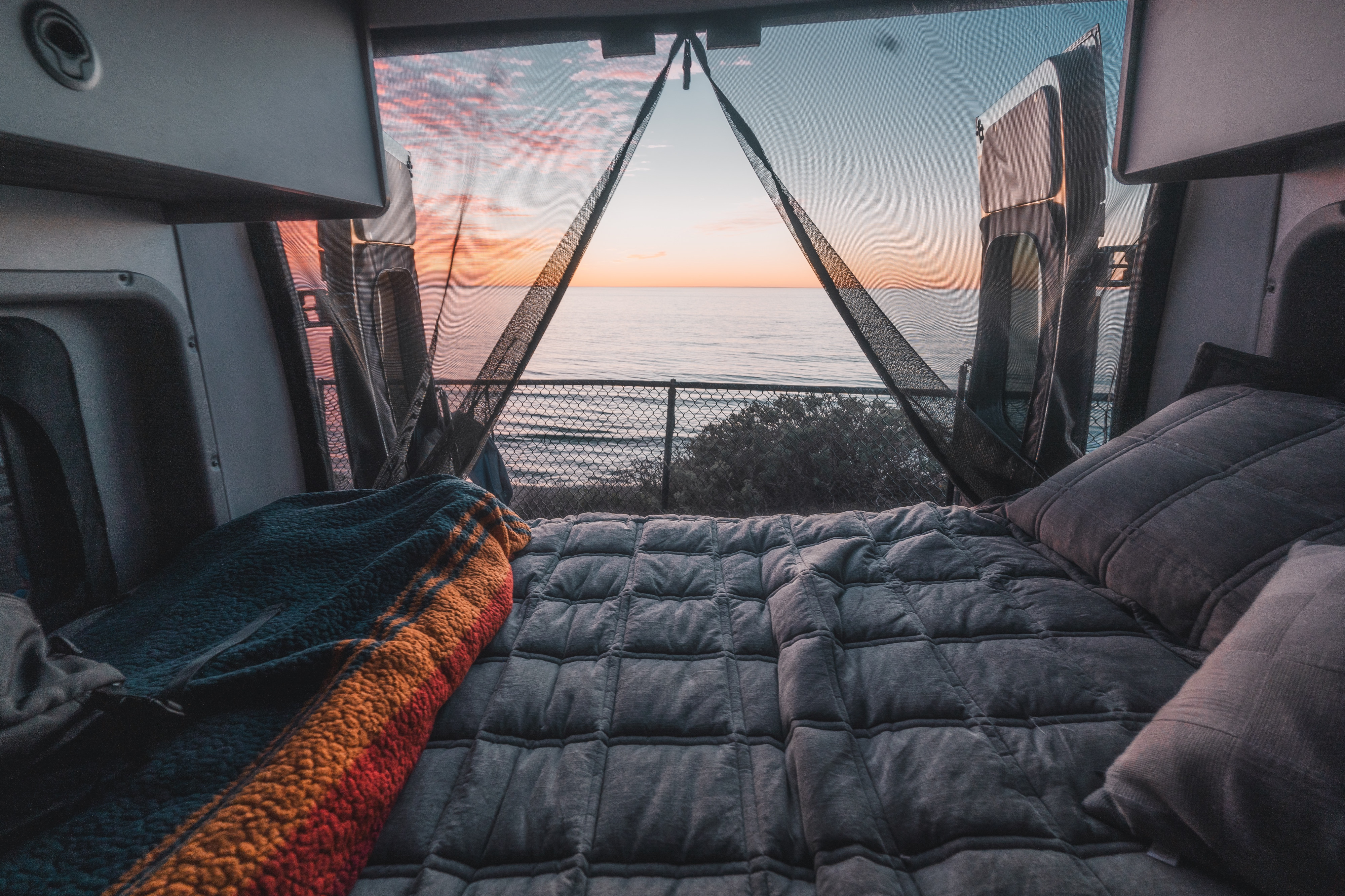 Cozy RV rental camper van bed facing an ocean sunset through screen-covered open doors, perfect for scenic retreats.. Winnebago Solis 2025