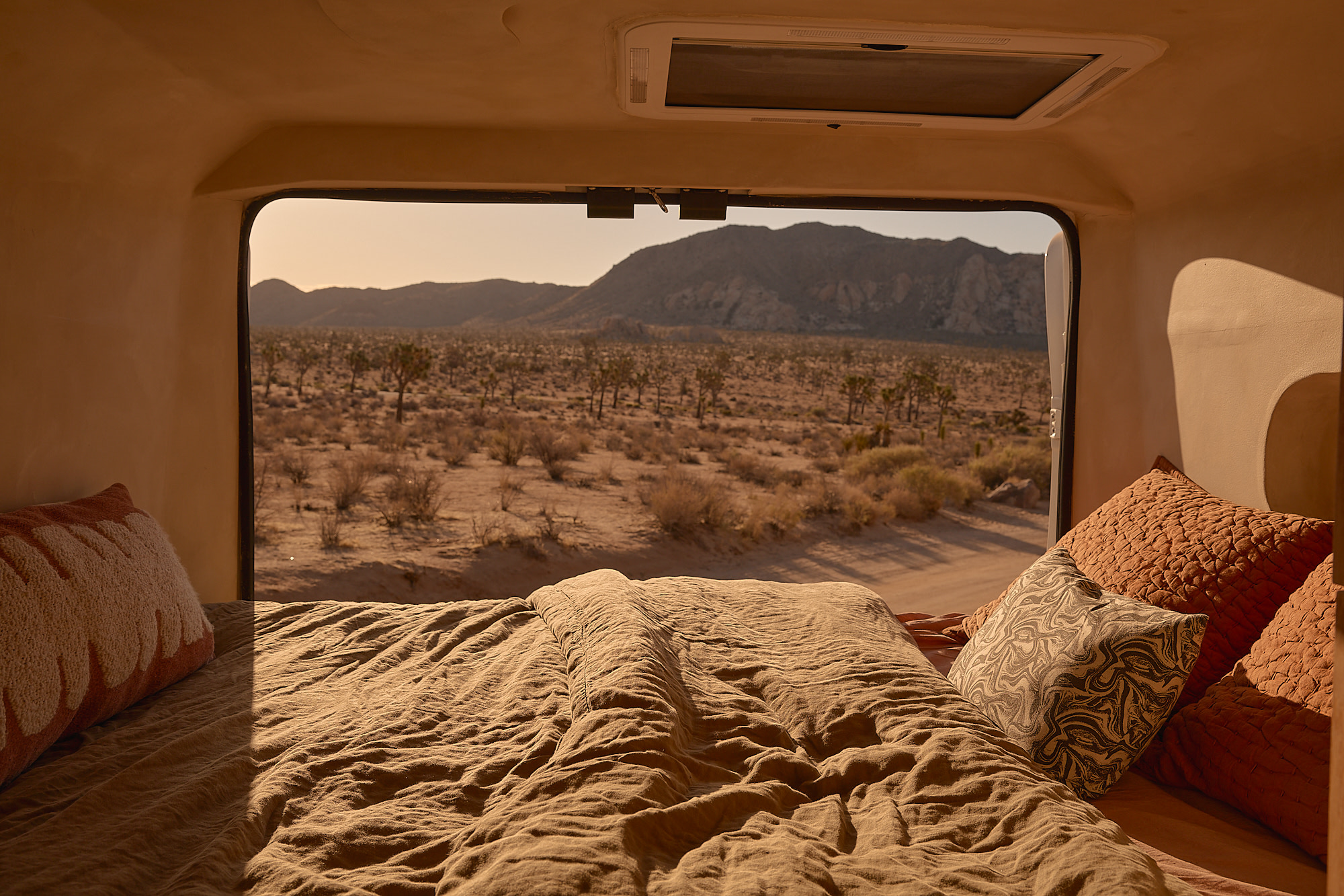 Inside an rv rental: a comfy bed with warm blankets, framing a window view of a desert, Joshua trees, & mountains.. Dodge Ram 1500 2021