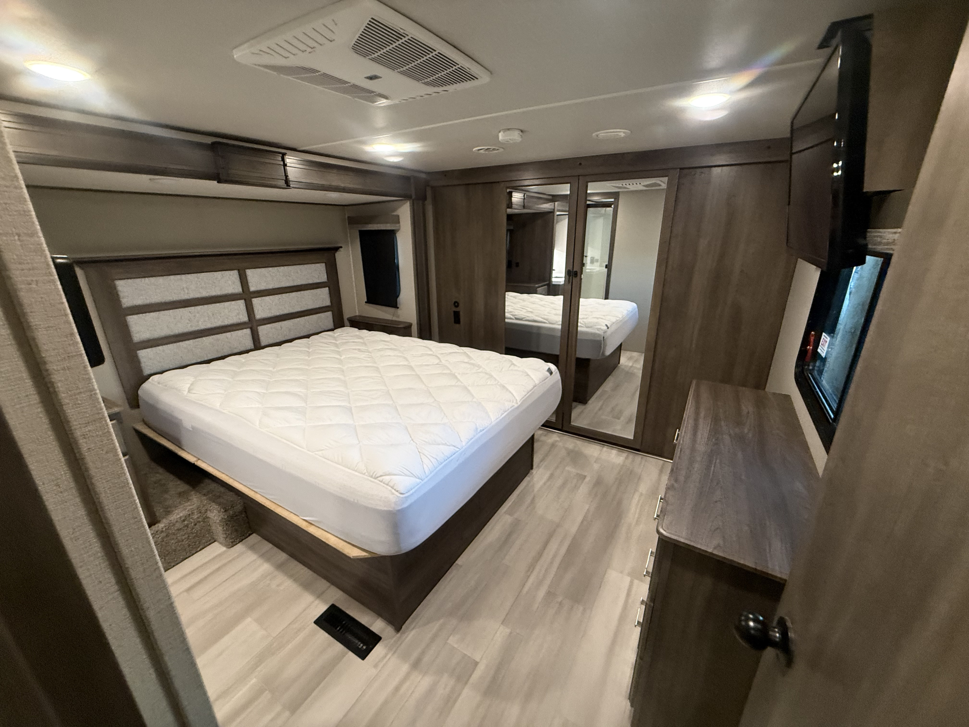 Bright RV rental bedroom features a large bed with quilted mattress, modern headboard, overhead AC, TV, and mirrored closet.. Grand Design Solitude S-Class 2021