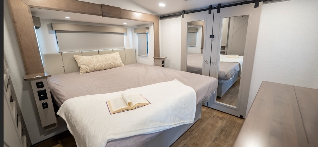 Modern RV rental bedroom with a large bed, open book on a white blanket, and mirrored sliding barn doors reflecting another room. Well-lit.. Alliance RV Avenue 2025