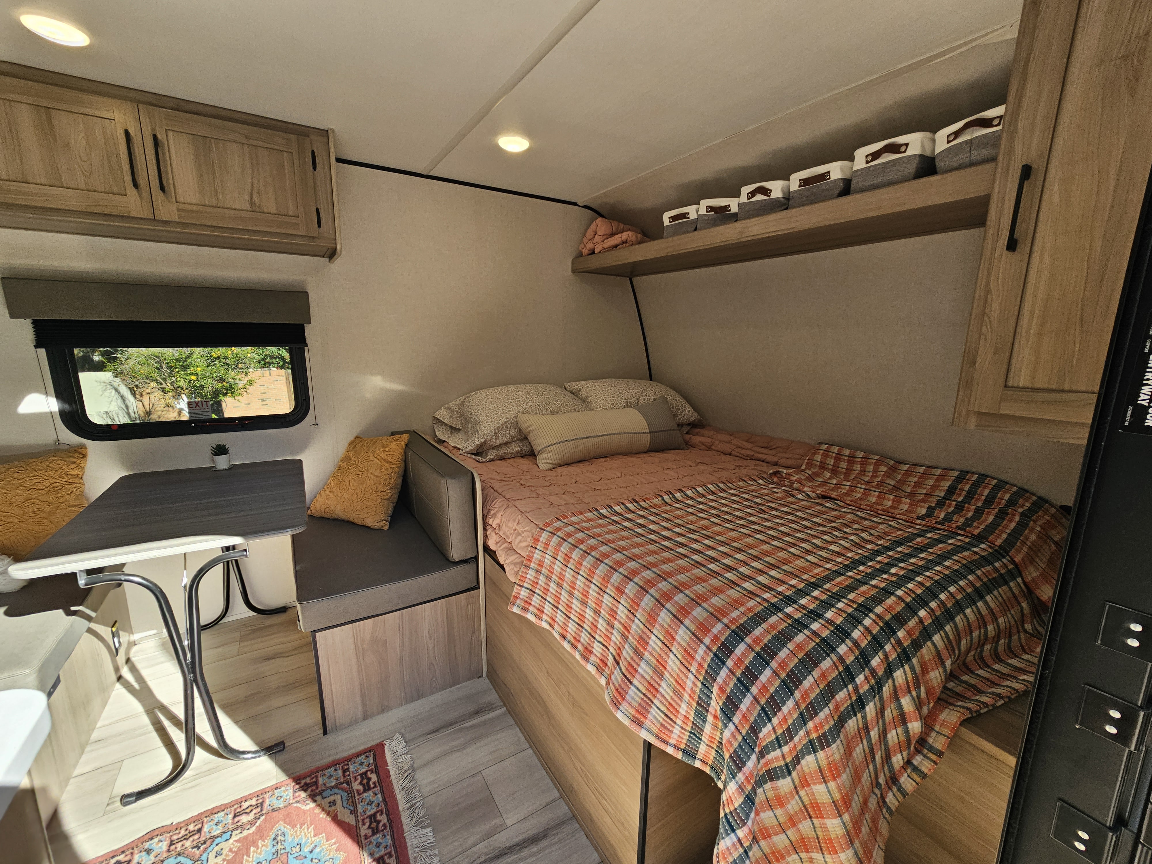 View inside a modern rv rental, showing a bed with a vibrant plaid blanket, a compact dinette, and light wood overhead cabinets.. Keystone Coleman 17BX 2025