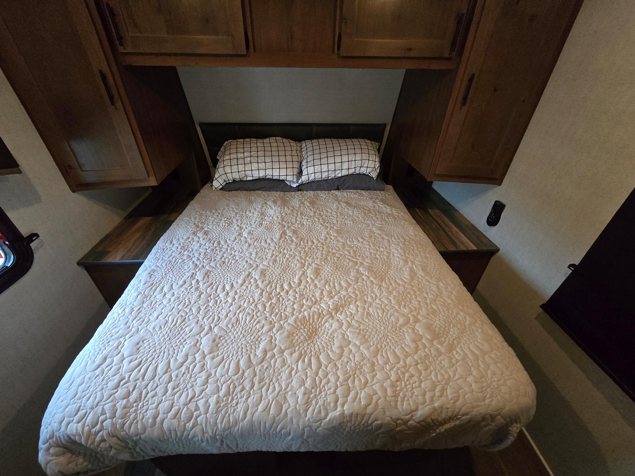 Cozy RV rental sleeping area: bed with quilted spread, grid pillows, dark headboard, and wooden cabinets overhead.. Outdoors RV Timber Ridge 2024