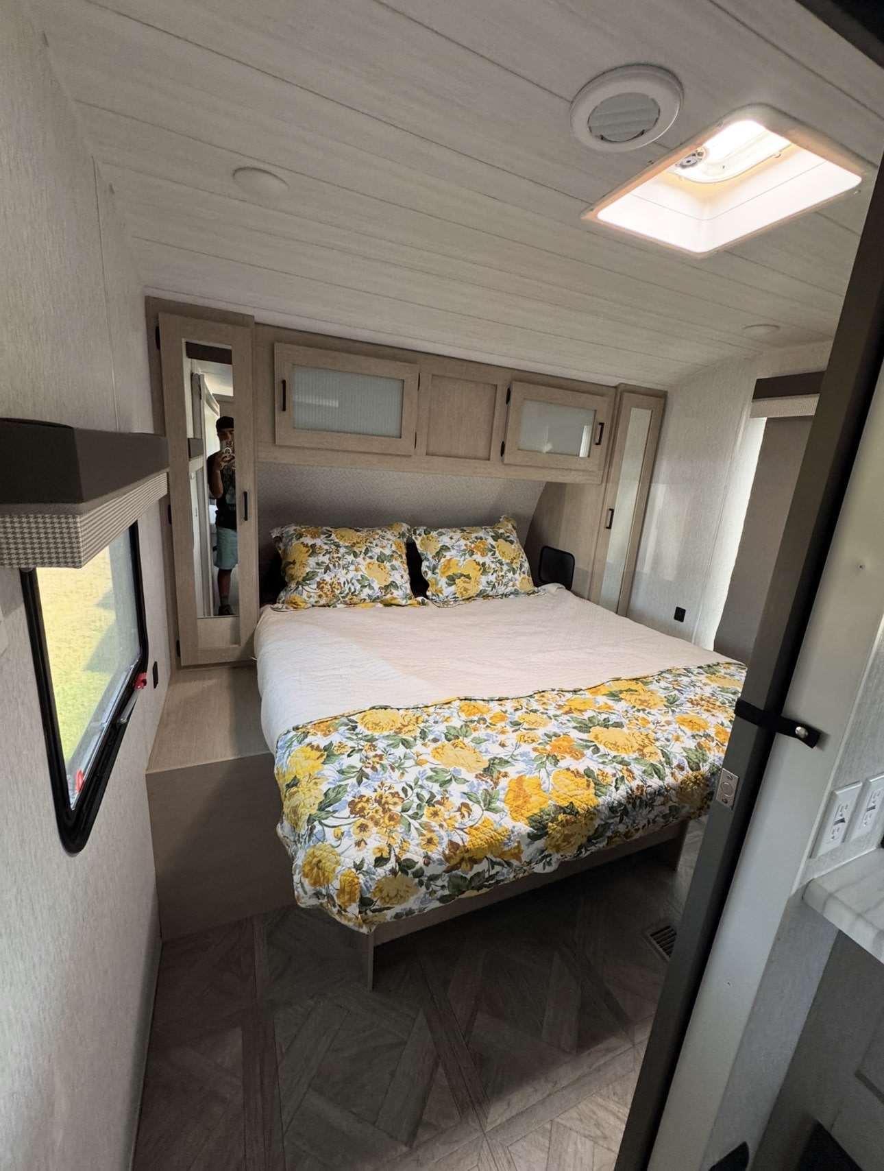 Cozy RV rental bedroom with a bed featuring yellow floral bedding, overhead cabinets, a window, and a mirror.. Forest River Wildwood 2023