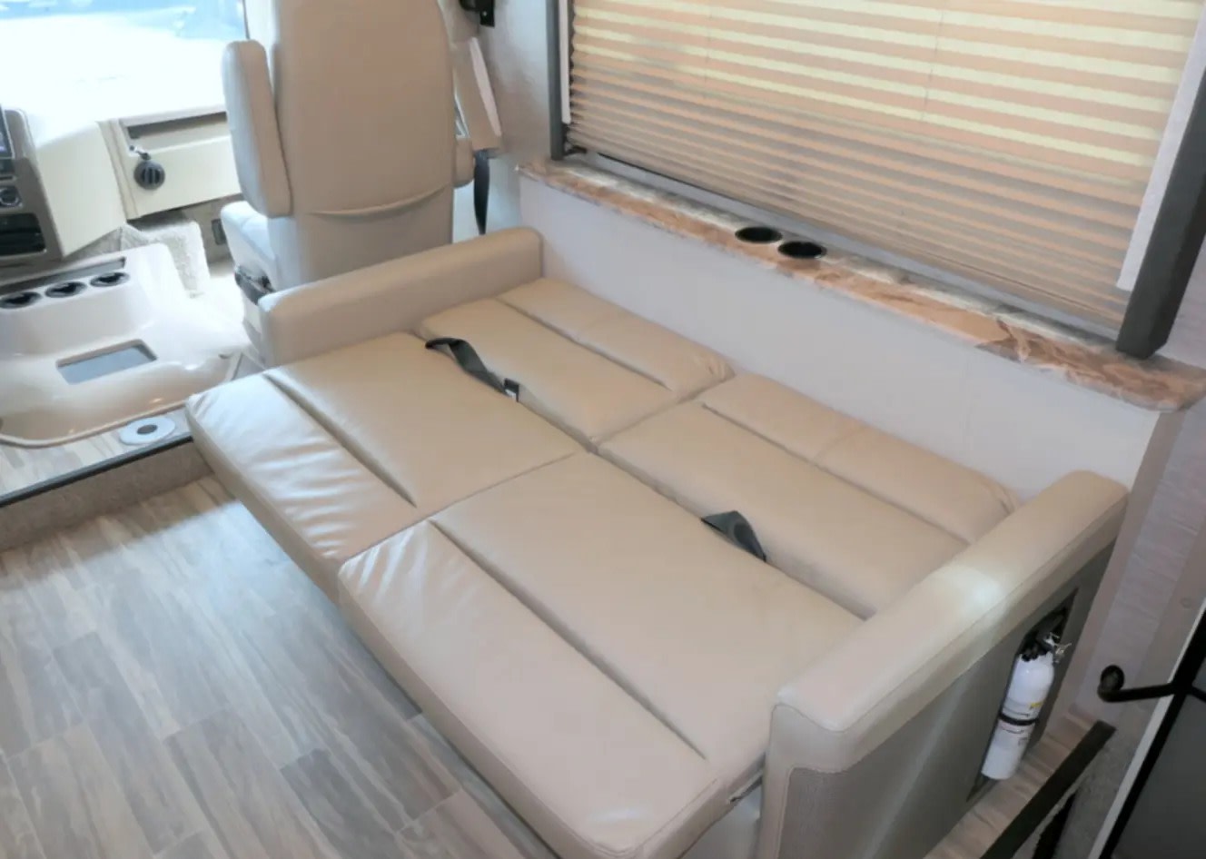 Inside an RV rental: a light beige sofa-bed is extended, facing the cockpit. Window blinds and a fire extinguisher are visible.. Thor A.C.E. 2022