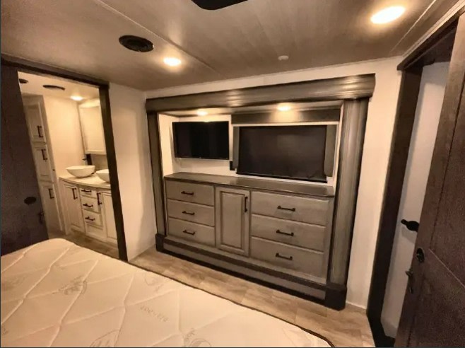Spacious RV rental bedroom features a dresser, dual TVs, and an ensuite bathroom entrance. Enjoy comfortable travel.. Keystone RV Montana 2023