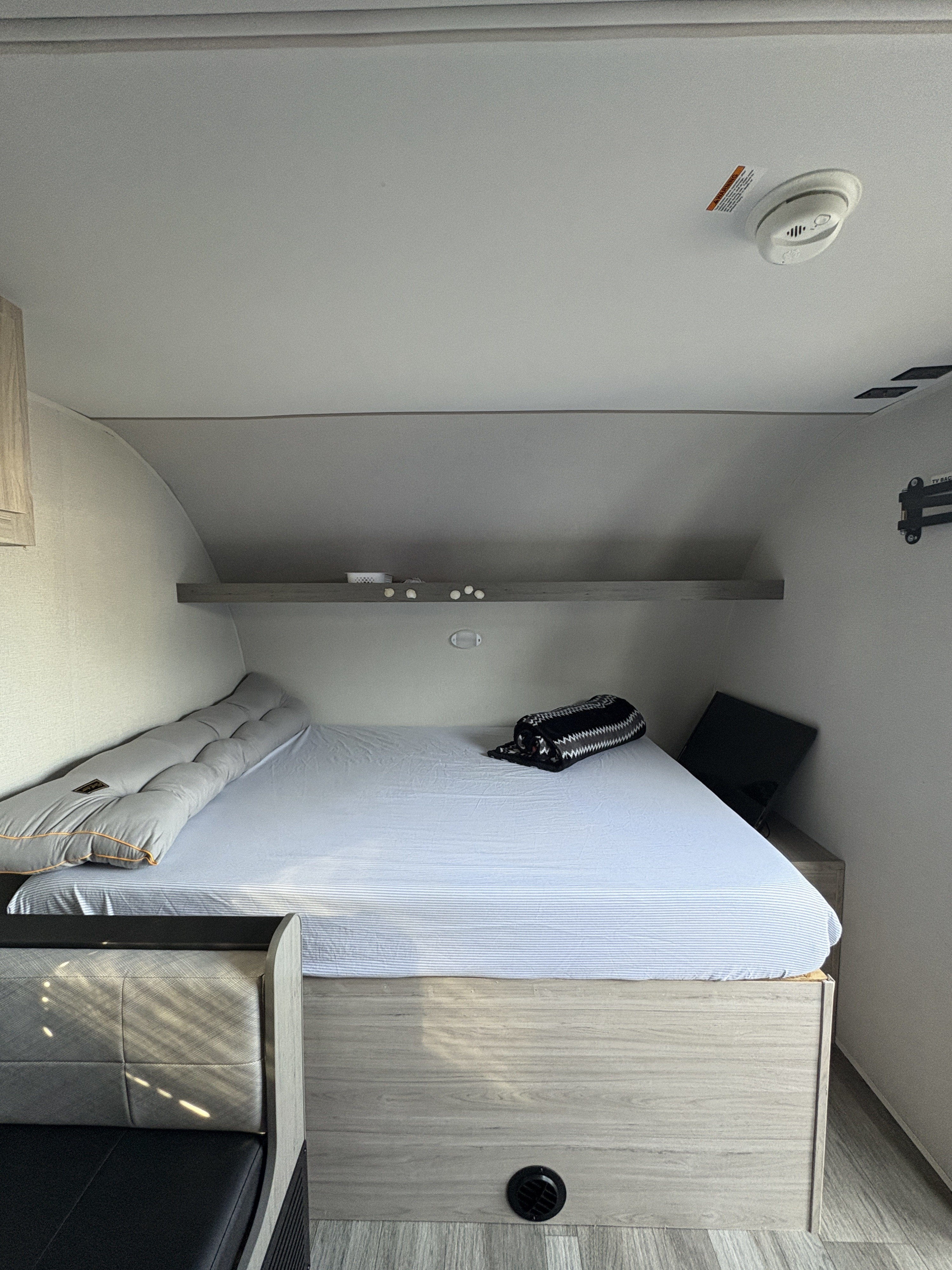 Cozy RV rental bedroom with a striped bed, bolster pillow, and a shelf. A smoke detector is on the ceiling and a TV mount on the wall.. Heartland RVs Mallard 2025