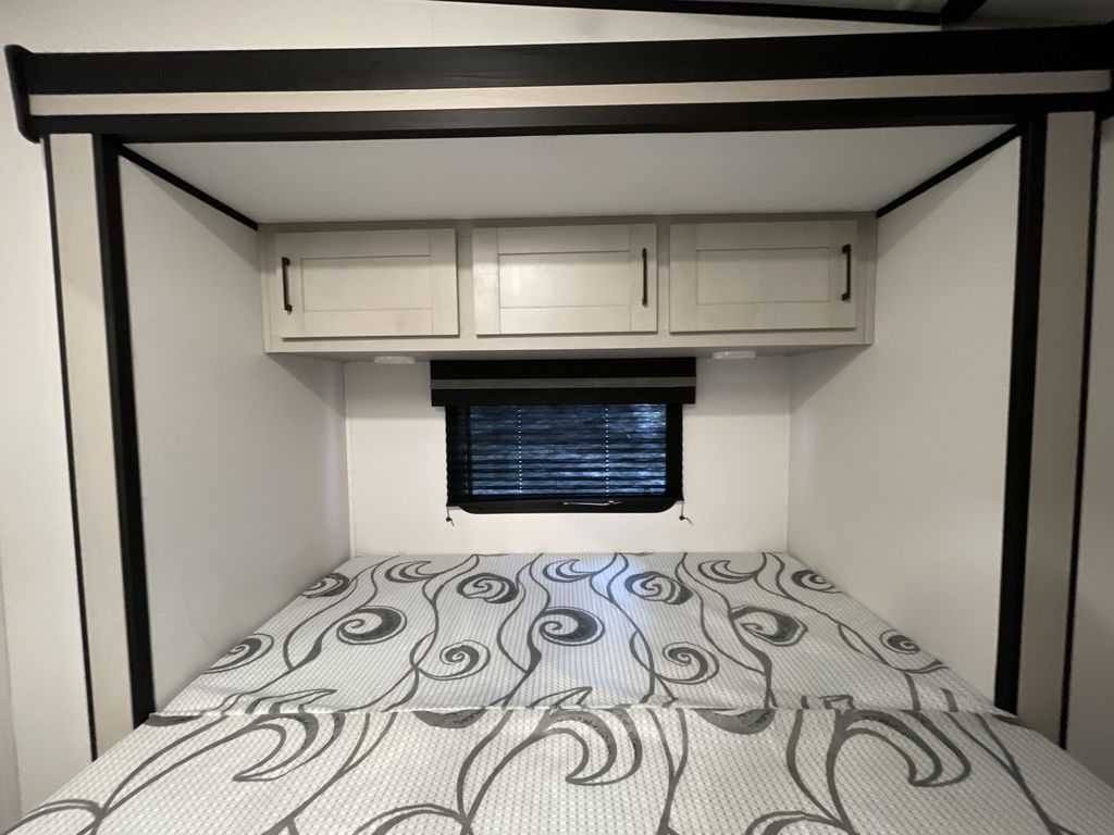 Cozy RV rental bedroom: patterned mattress, overhead storage cabinets, and a window with blinds, framed by black trim.. Heartland Torque 2022