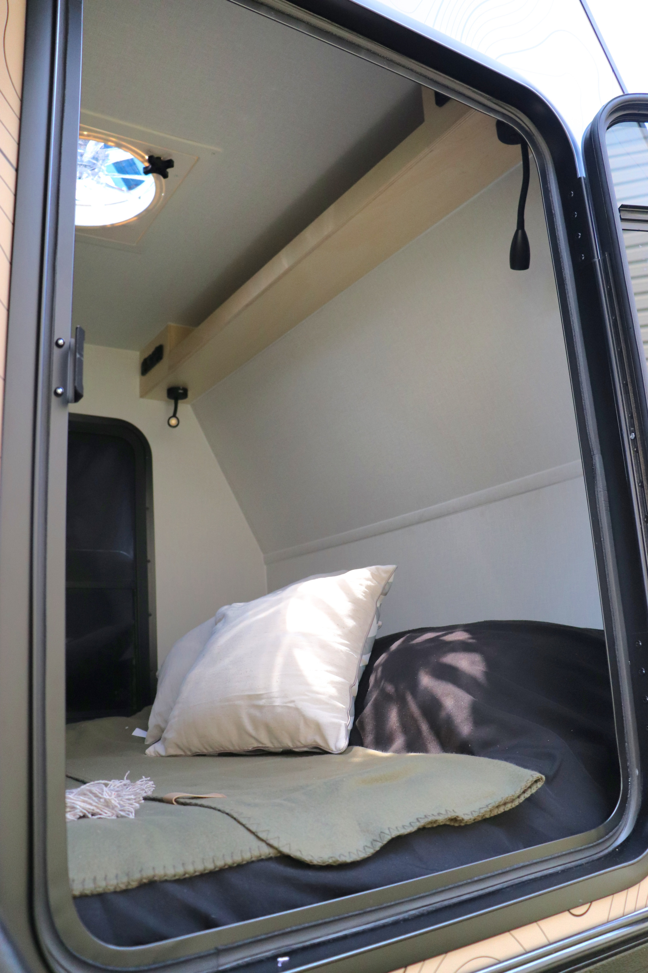 Peek inside a cozy RV rental bedroom. Features a comfortable bed with a white pillow and a green blanket, ready for your next adventure.. Mud Overland Trailer Québec Xpédition 2025