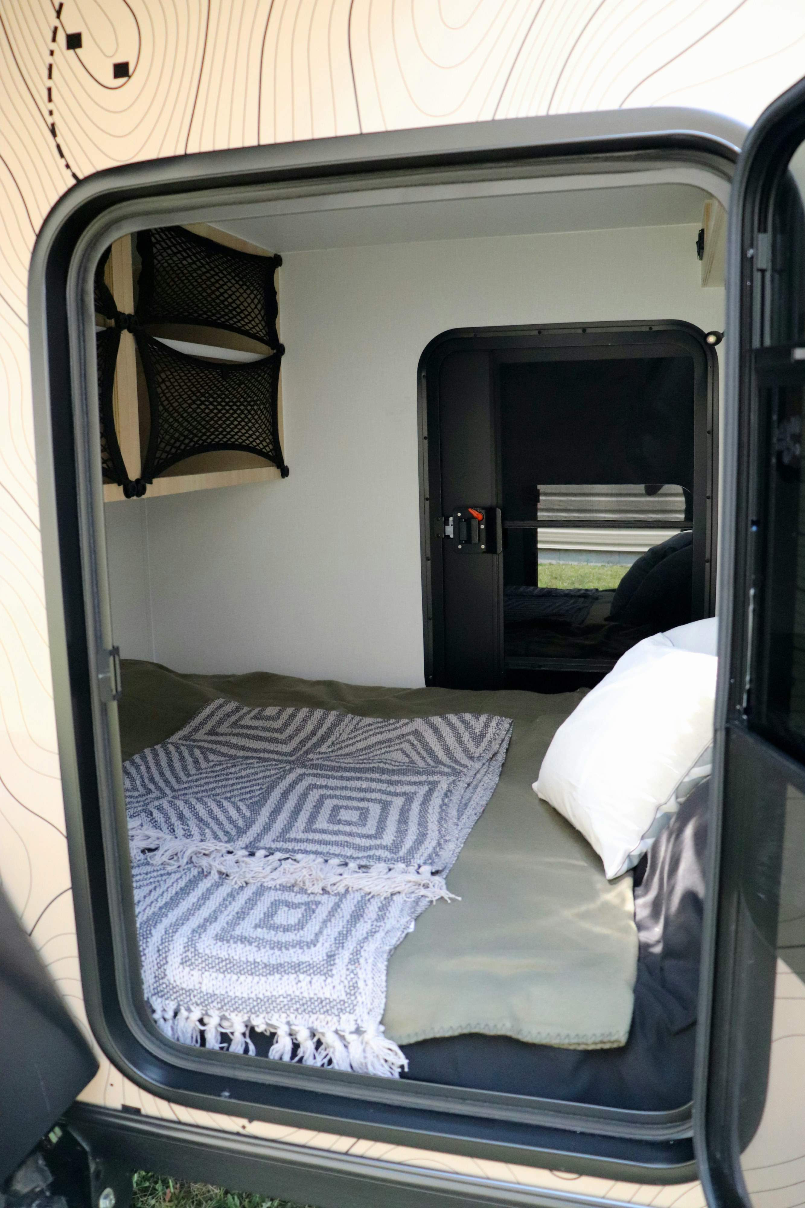 This cozy rv rental features a compact camper interior. A bed with a patterned blanket, pillow, and mesh storage nets are visible.. Mud Overland Trailer Québec Xpédition 2025