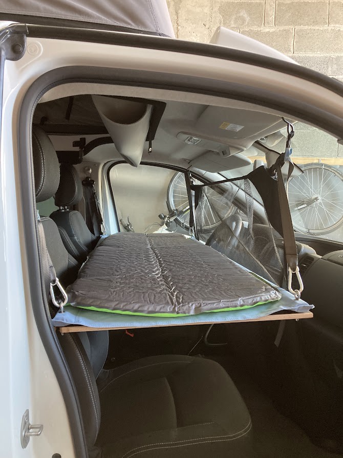 Compact camper van setup: a gray sleeping pad on a wood platform extends over the passenger seat, with overhead storage for rv rental travel.. Renault Trafic 2025