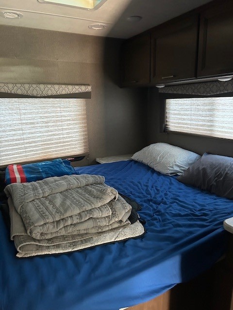 Interior of an rv rental bedroom with a bed, blue sheets, pillows, folded blankets, and overhead cabinets.. Thor Freedom Elite 2021
