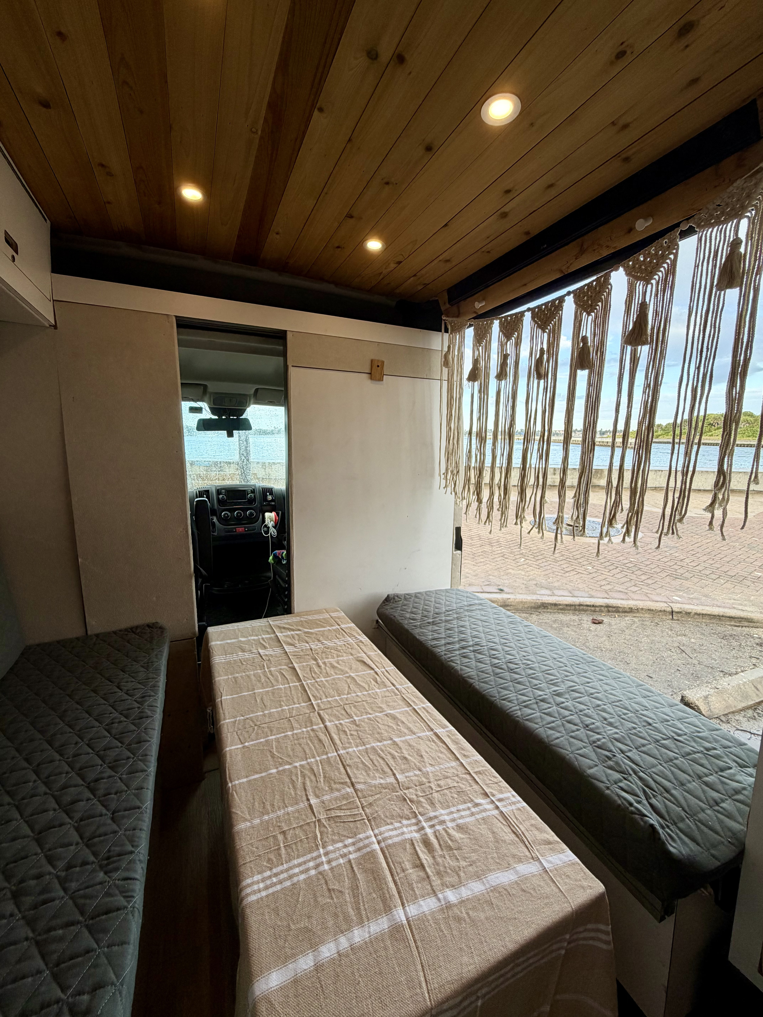 Cozy RV rental interior with wooden ceiling, quilted beds/benches, macrame curtain, and a scenic waterfront view outside.. Gmc Ram Promaster 2018