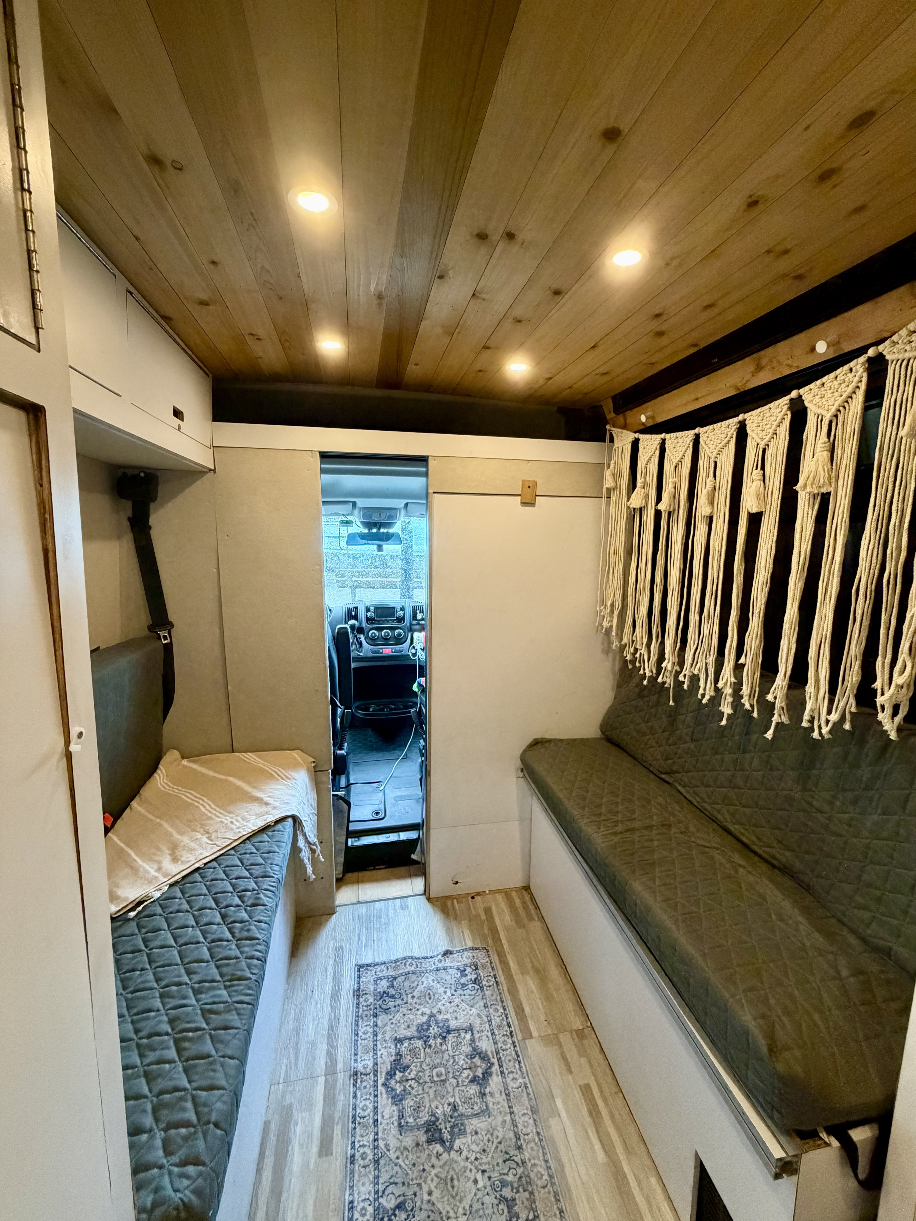 Cozy RV rental camper van interior. Features a bright wood plank ceiling with lights, cushioned benches, and a boho macrame wall hanging.. Gmc Ram Promaster 2018