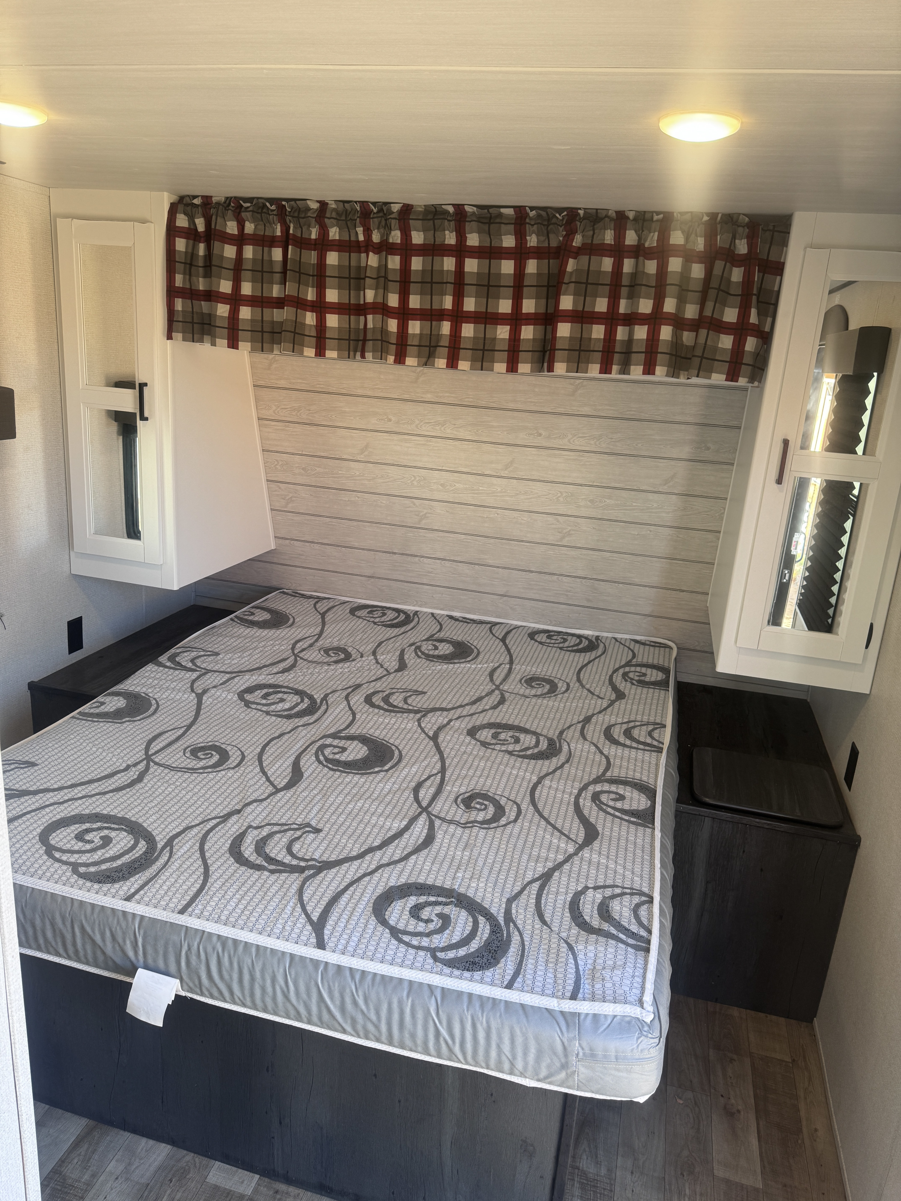 Inside an RV rental: a compact bedroom with a patterned bed, storage cabinets, plaid curtains, and wood paneling.. Keystone RV Hideout 2022